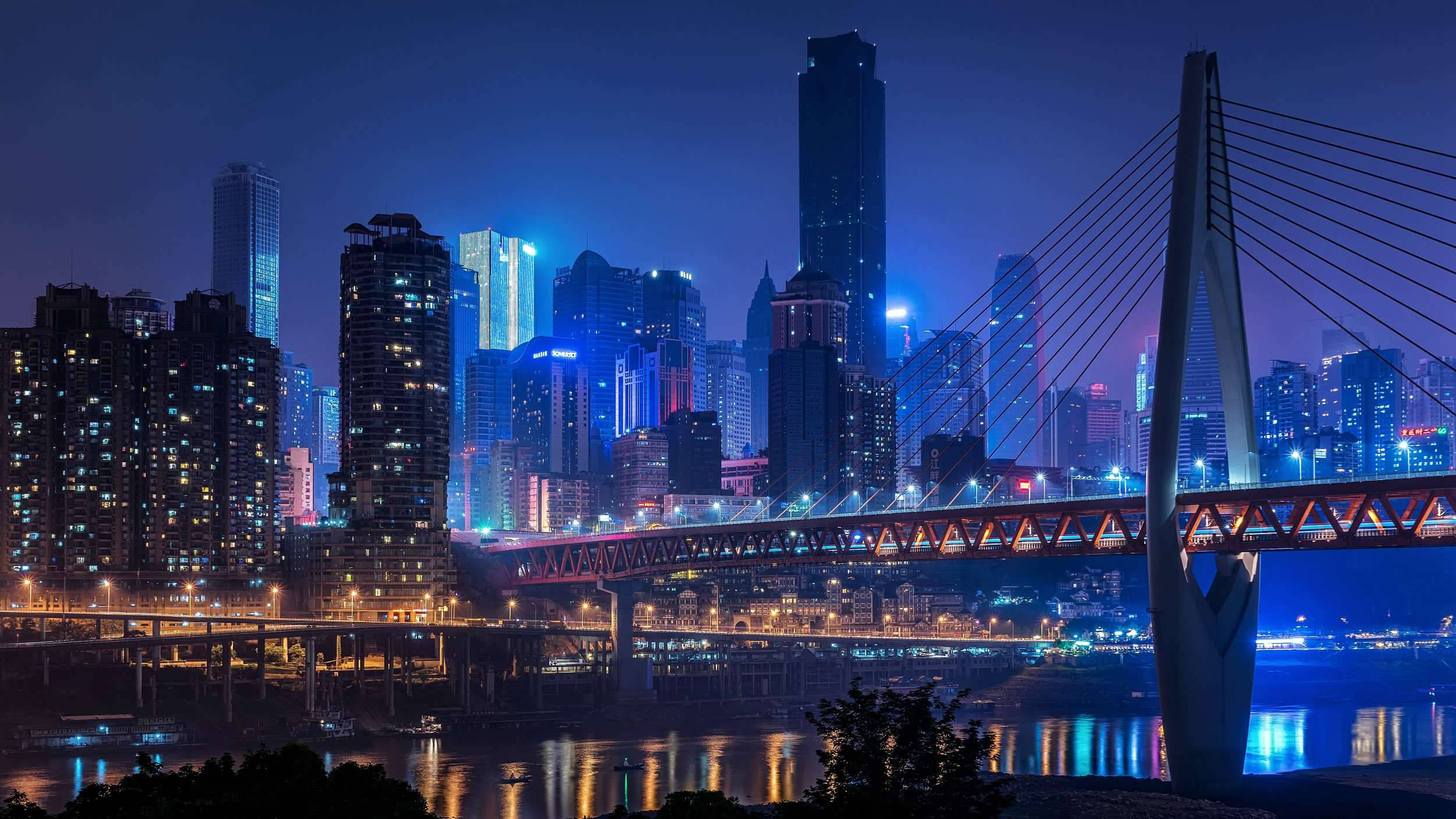Chongqing 桌布 壁紙 — 充滿活力的夜景城市景觀，一座發光的橋樑橫跨河流，反射著高聳摩天大樓的霓虹燈光。