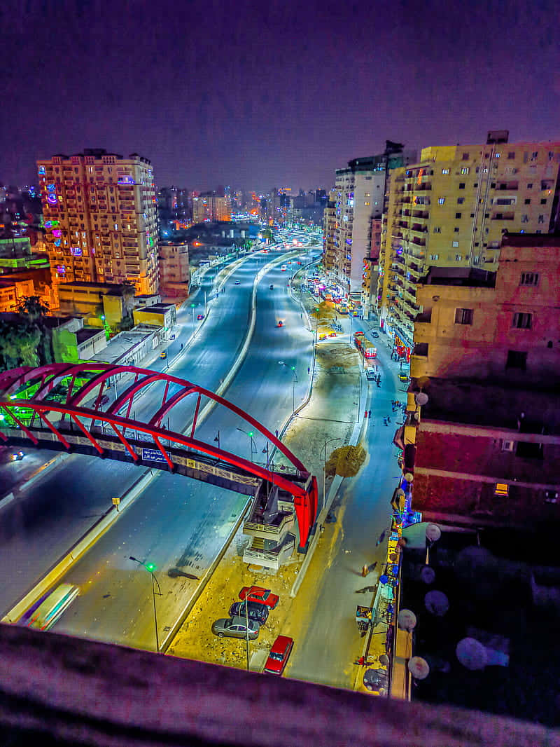 亞歷山卓 桌布 壁紙 — 充滿活力的夜景城市景觀，一座發光的紅色橋樑拱形橫跨在繁忙的高速公路上，兩旁是燈火通明的高層建築。