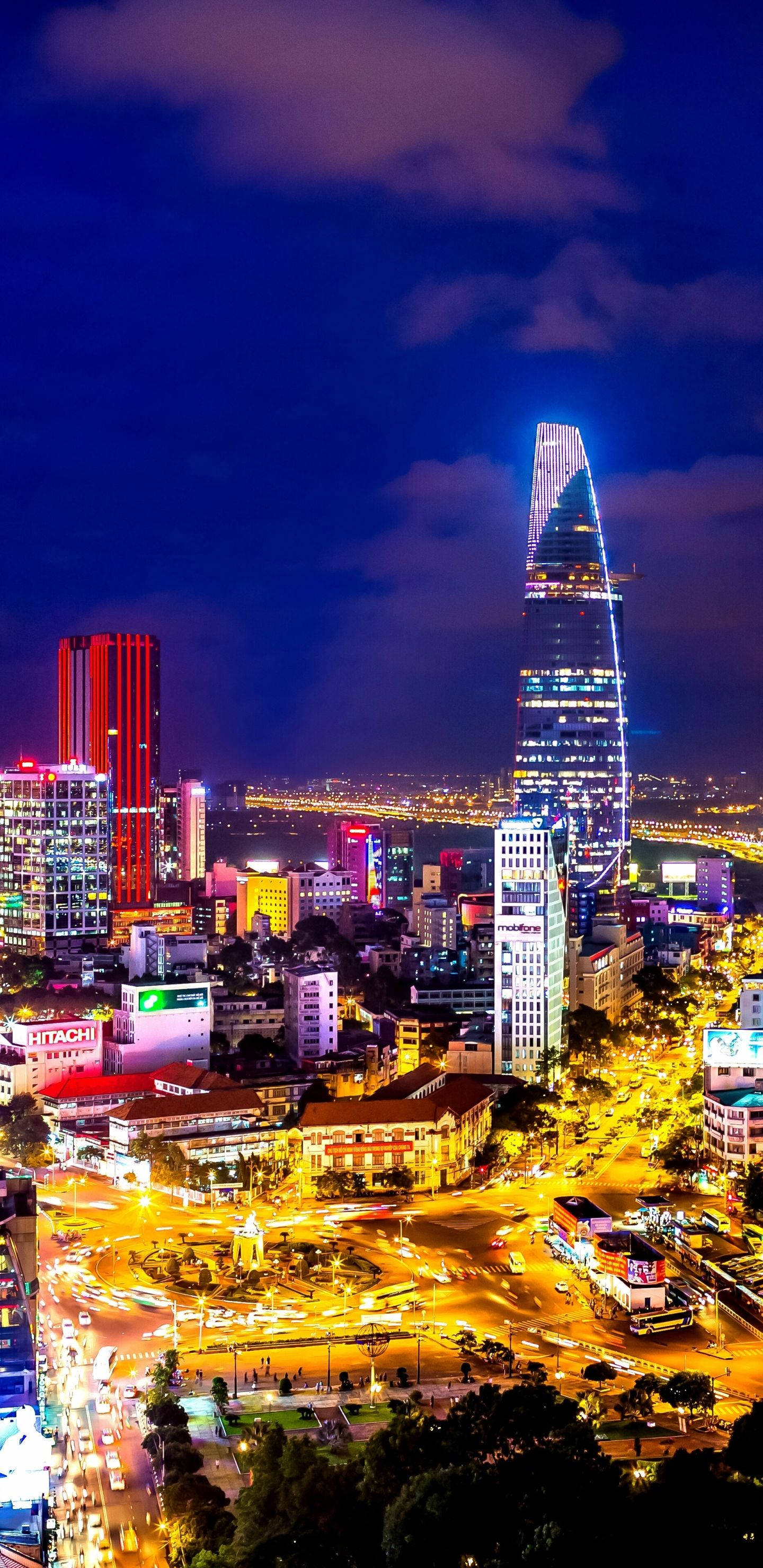 胡志明市 桌布 壁紙 — 充滿活力的夜景城市景觀，一座高聳的摩天大樓在深紫色天空和下方繁忙街道的映襯下發出藍色光芒。