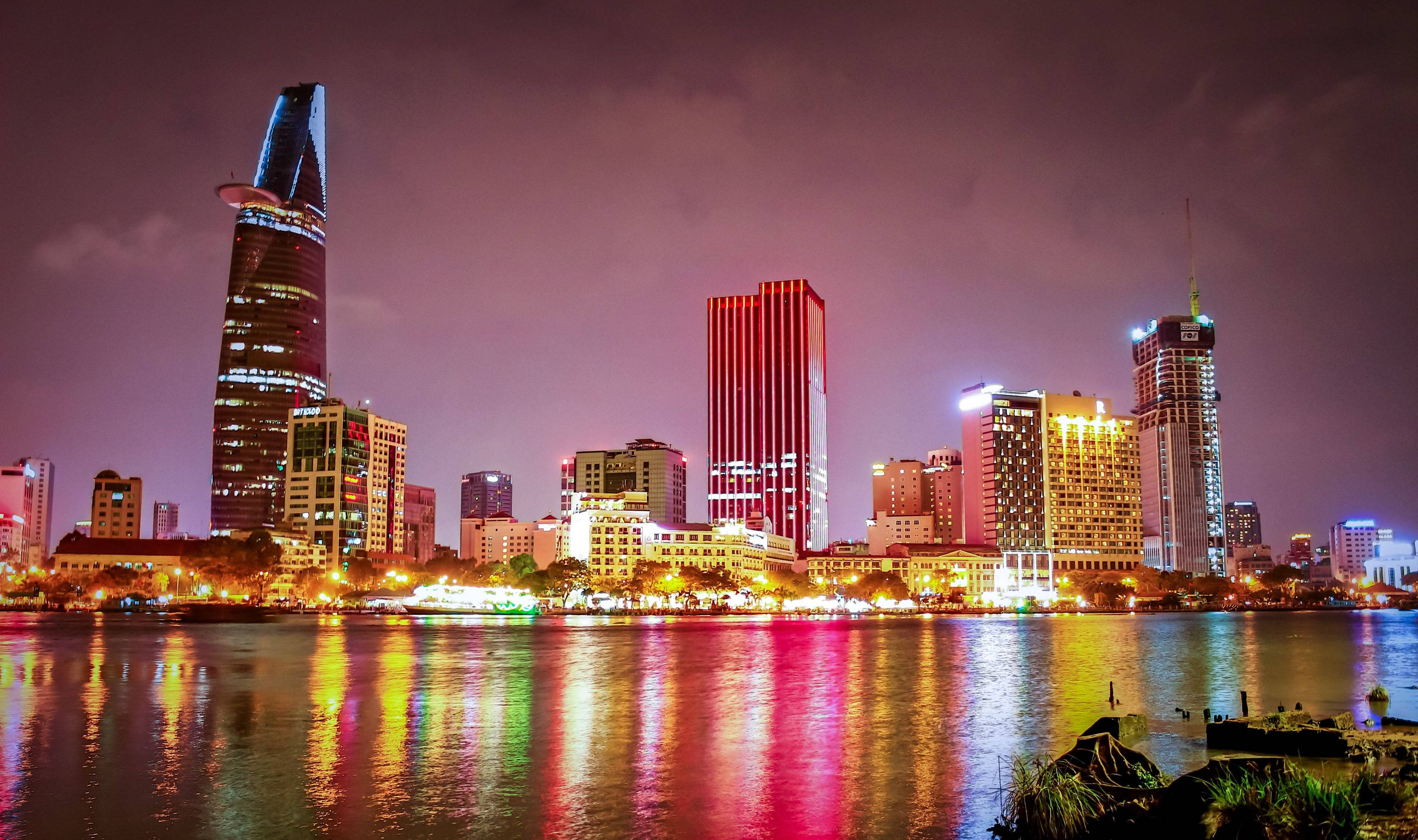 Ho Chi Minh City 桌布 壁紙 — 充滿活力的夜景城市景觀，在昏暗的紫色天空下，寧靜的河流中映照著燈火通明的摩天大樓。
