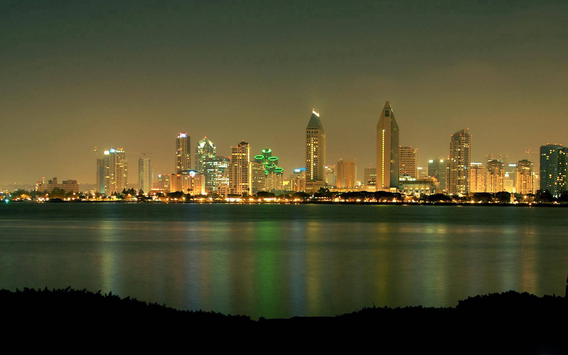 Sans 桌布 壁紙 — 充滿活力的夜景城市景觀，在朦朧的天空下，燈火通明的摩天大樓倒映在平靜的水面上。