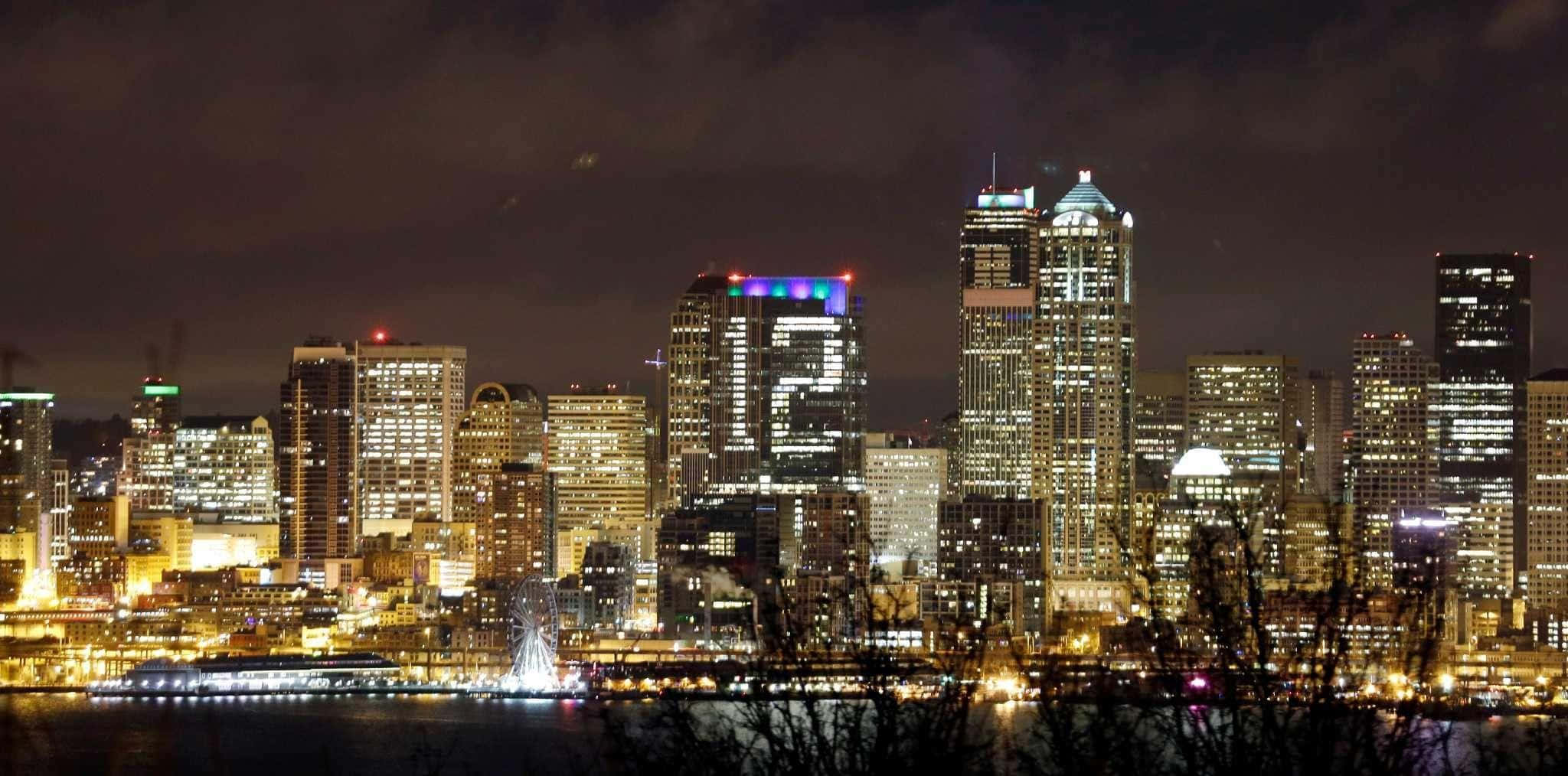 Seattle At Night 桌布 壁紙 — 充滿活力的夜景城市景觀，在黑暗多雲的天空下，燈火通明的摩天大樓倒映在水面上。