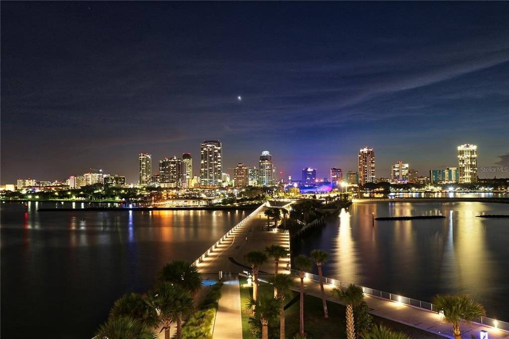 Evening 桌布 壁紙 — 充滿活力的夜景城市景觀，明亮的摩天大樓倒映在平靜的水面上，由一條燈火通明、兩旁種滿棕櫚樹的步道連接。
