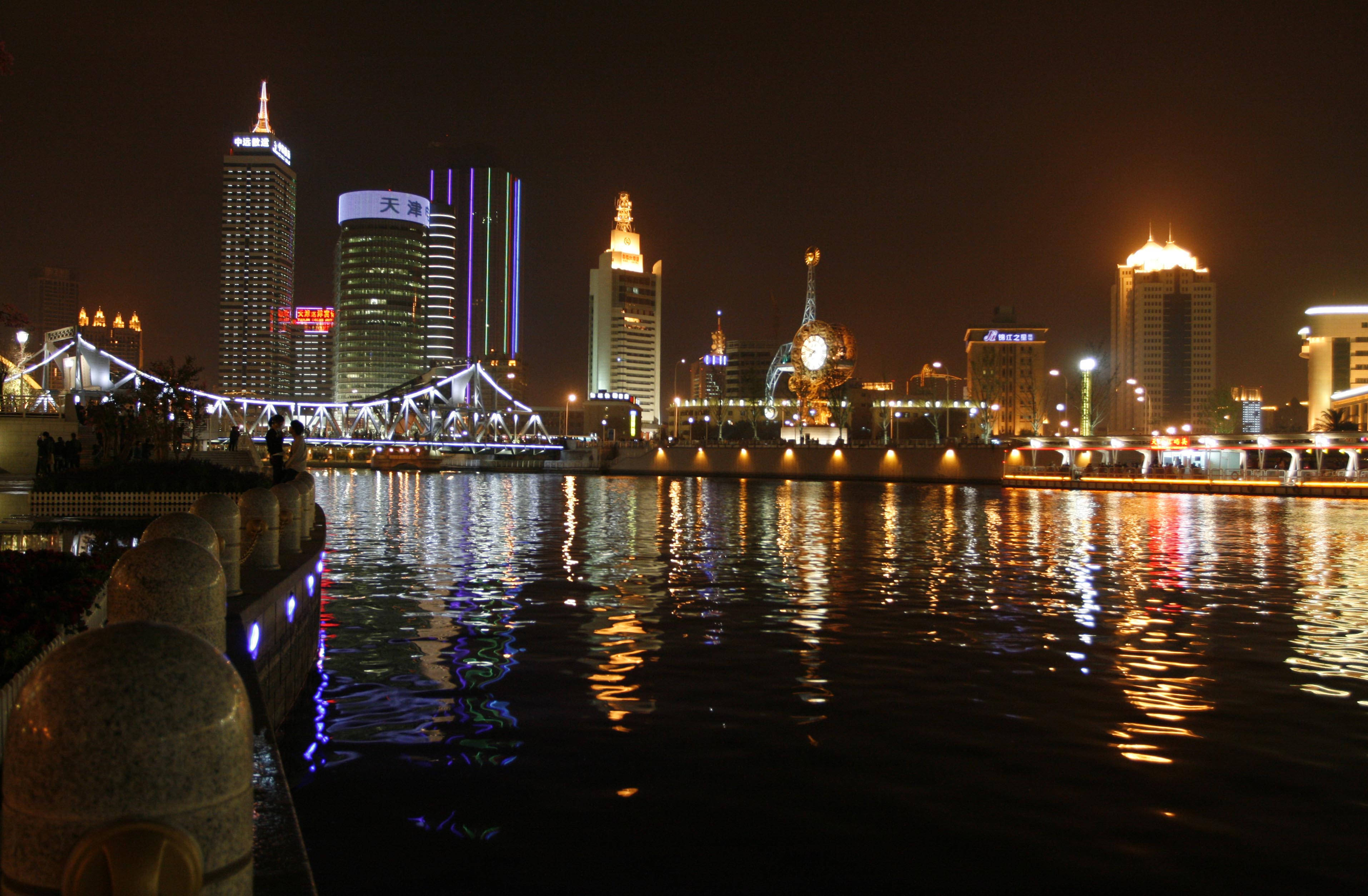 Tianjin 桌布 壁紙 — 充滿活力的夜景城市景觀，有燈火通明的摩天大樓和發光的橋樑倒映在黑暗的水面上。