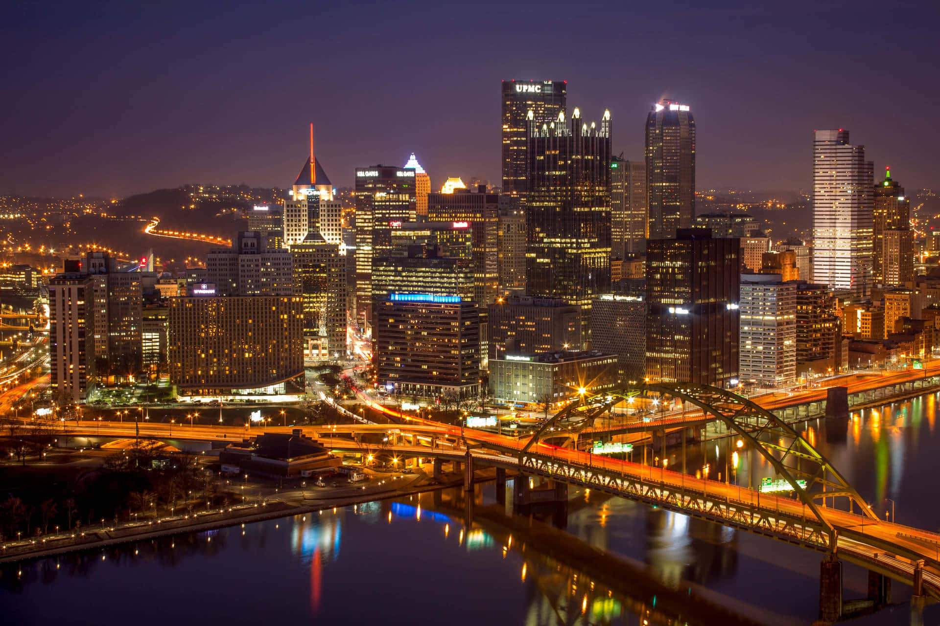 Pittsburgh Skyline 桌布 壁紙 — 充滿活力的夜景城市景觀，有燈火通明的摩天大樓和一座在黑暗河流中閃爍倒影的橋樑。