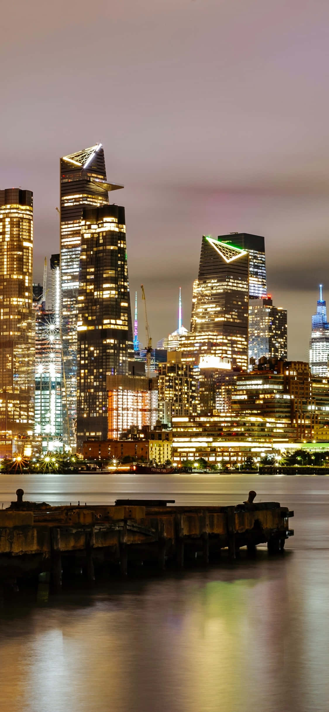 iPhone Xs New York 桌布 壁紙 — 充滿活力的夜景城市景觀，燈火通明的摩天大樓倒映在平靜的水面上，前景是飽經風霜的碼頭。