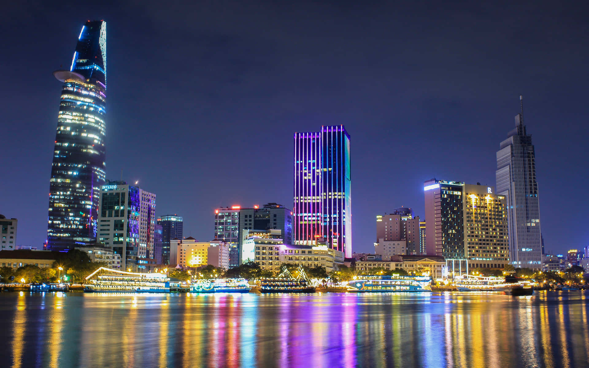 越南 桌布 壁紙 — 充滿活力的夜景城市景觀，燈火通明的摩天大樓倒映在下方平靜的水面上。