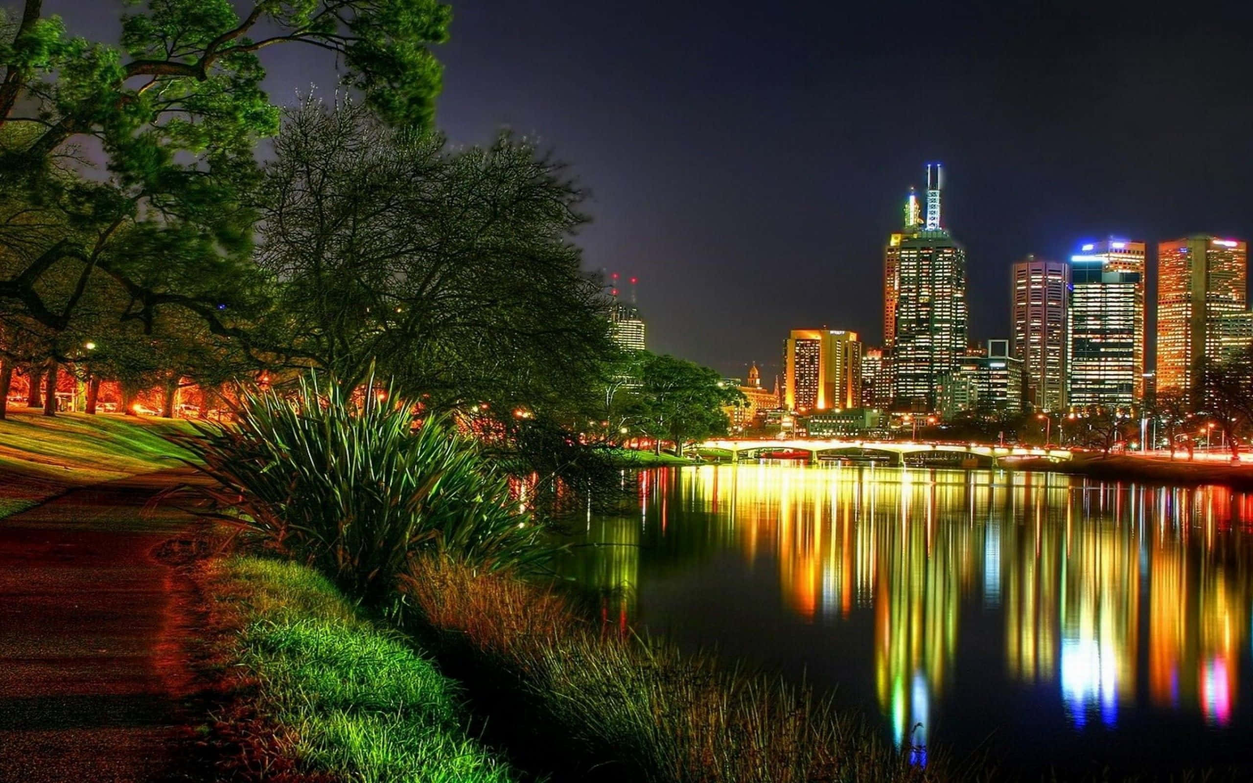 Beautiful City 桌布 壁紙 — 充滿活力的夜景城市景觀，燈火通明的摩天大樓倒映在平靜的河面上，兩旁是茂密的綠樹和蜿蜒的小徑。