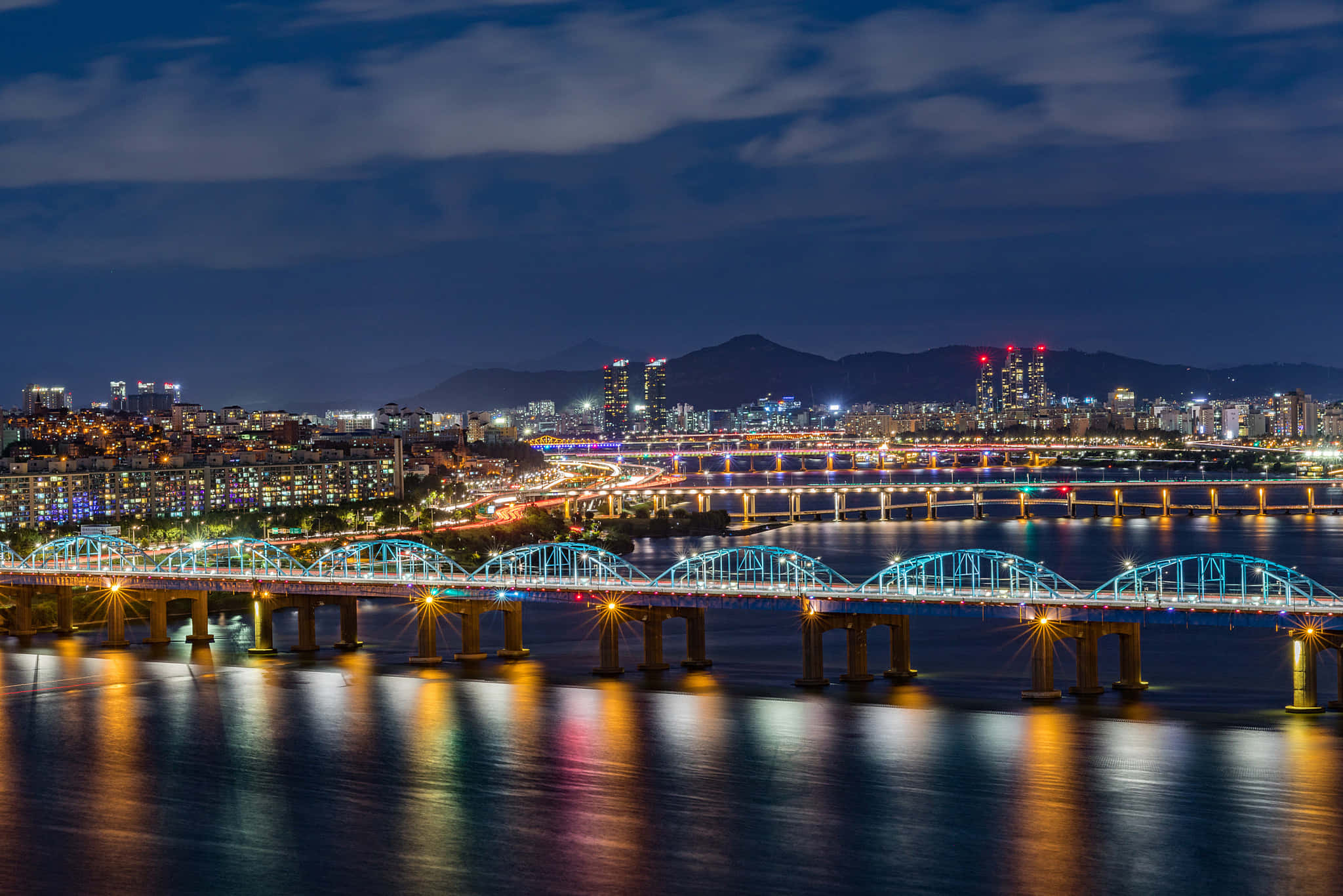 南韓 桌布 壁紙 — 充滿活力的夜景城市景觀，特色是水面上燈火通明的橋樑，發光的燈光在水面反射，遠處的山脈在黑暗的天空下。