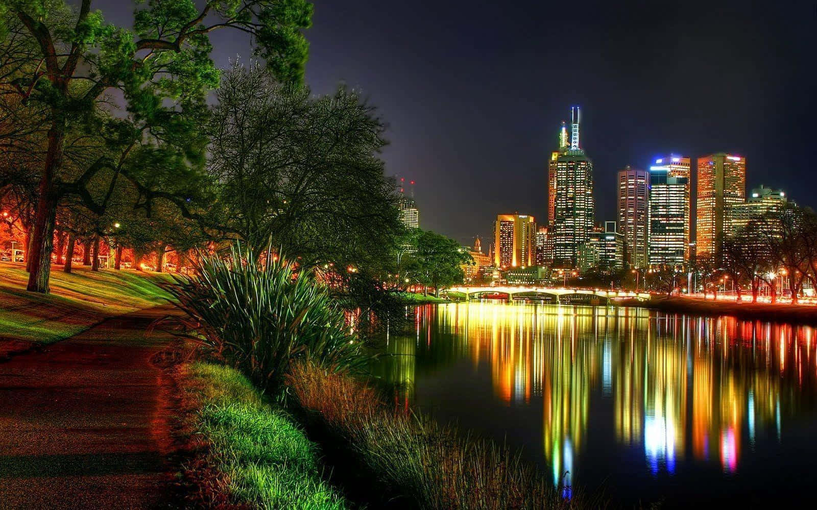 Scenic Desktop 桌布 壁紙 — 充滿活力的夜景城市景觀，發光的摩天大樓倒映在平靜的河流中，兩旁是燈火通明的樹木和小徑。