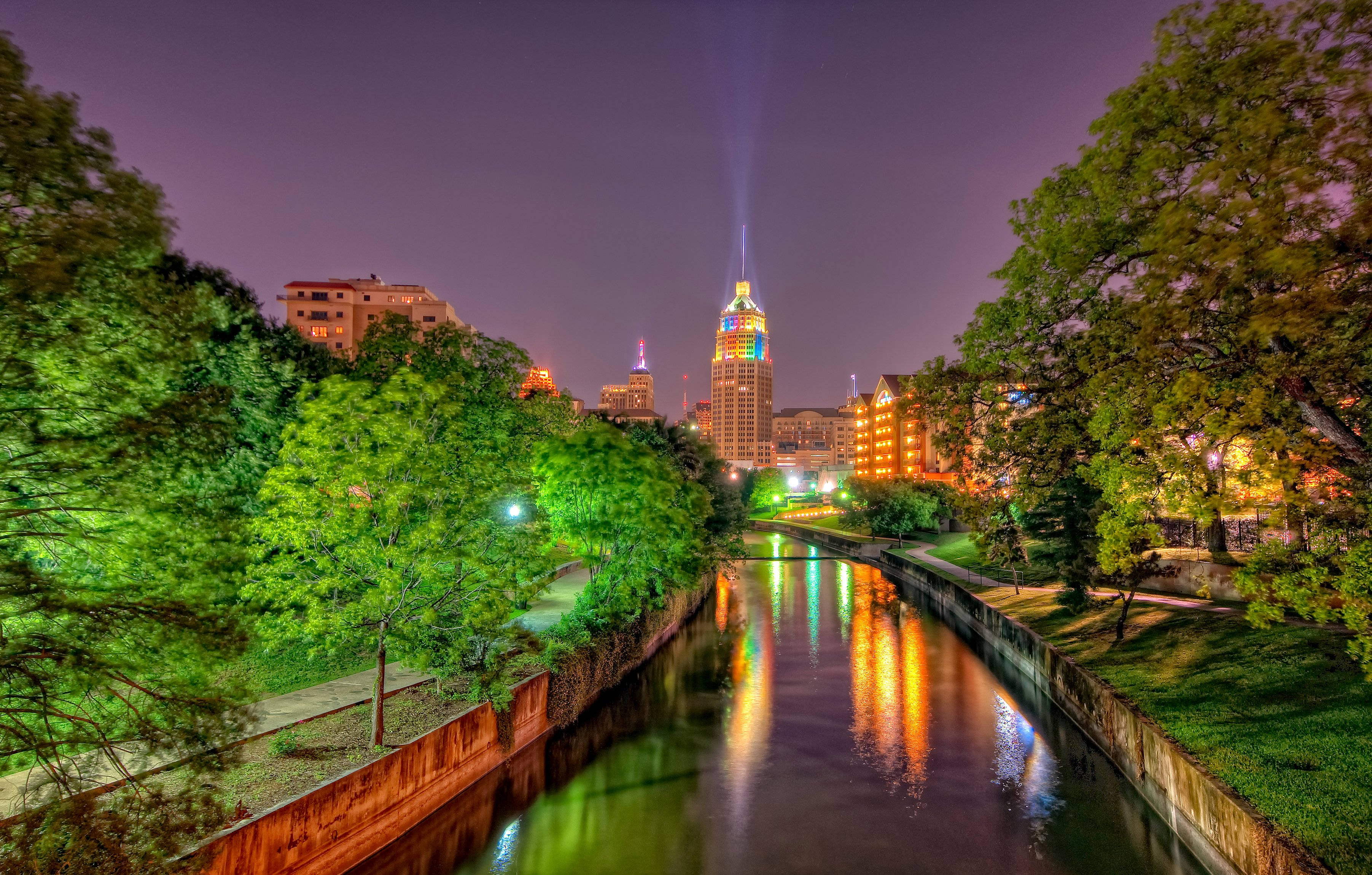 San Antonio 桌布 壁紙 — 充滿活力的夜景城市景觀，發光的摩天大樓倒映在平靜的河流中，兩旁是茂密的綠樹。