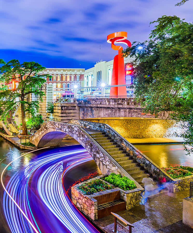 San Antonio 桌布 壁紙 — 充滿活力的夜景城市景觀，石橋橫跨運河，運河上有來往車輛留下的藍色和紫色光軌。