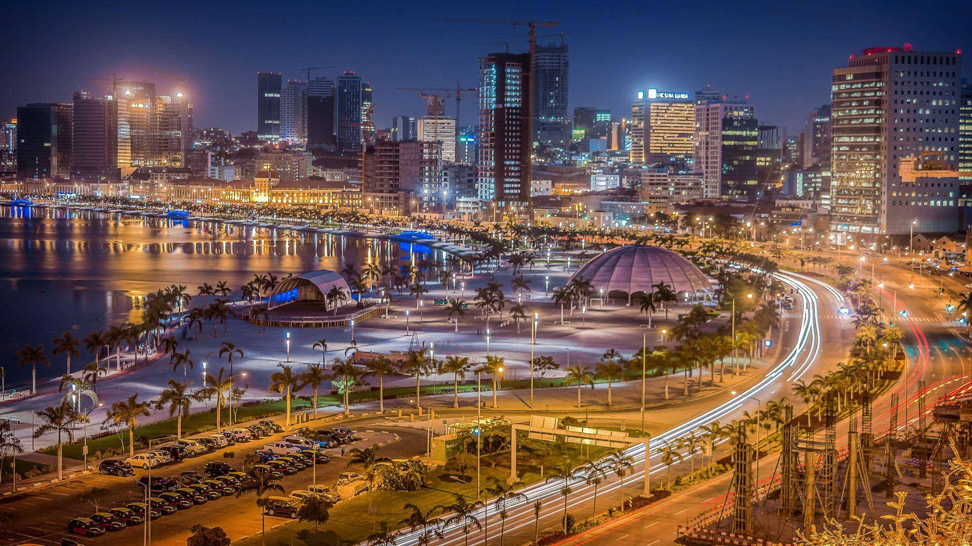 Angola 桌布 壁紙 — 充滿活力的夜景城市景觀，閃爍的摩天大樓倒映在水面上，繁忙道路上的燈光形成光帶。