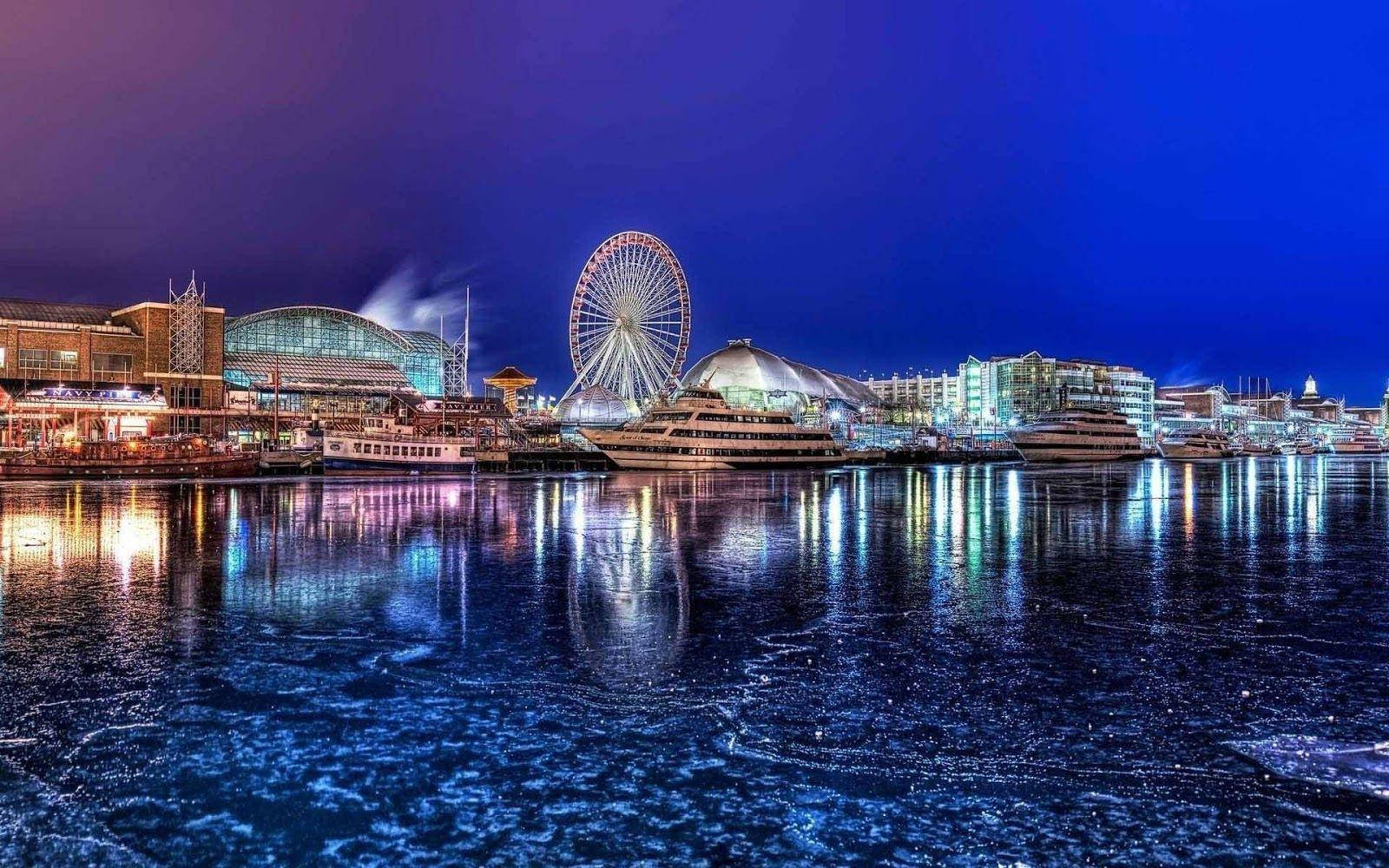 Night Aesthetic 桌布 壁紙 — 充滿活力的夜景城市景觀，閃爍的摩天輪和現代建築倒映在冰冷的水中，深藍色的天空下。
