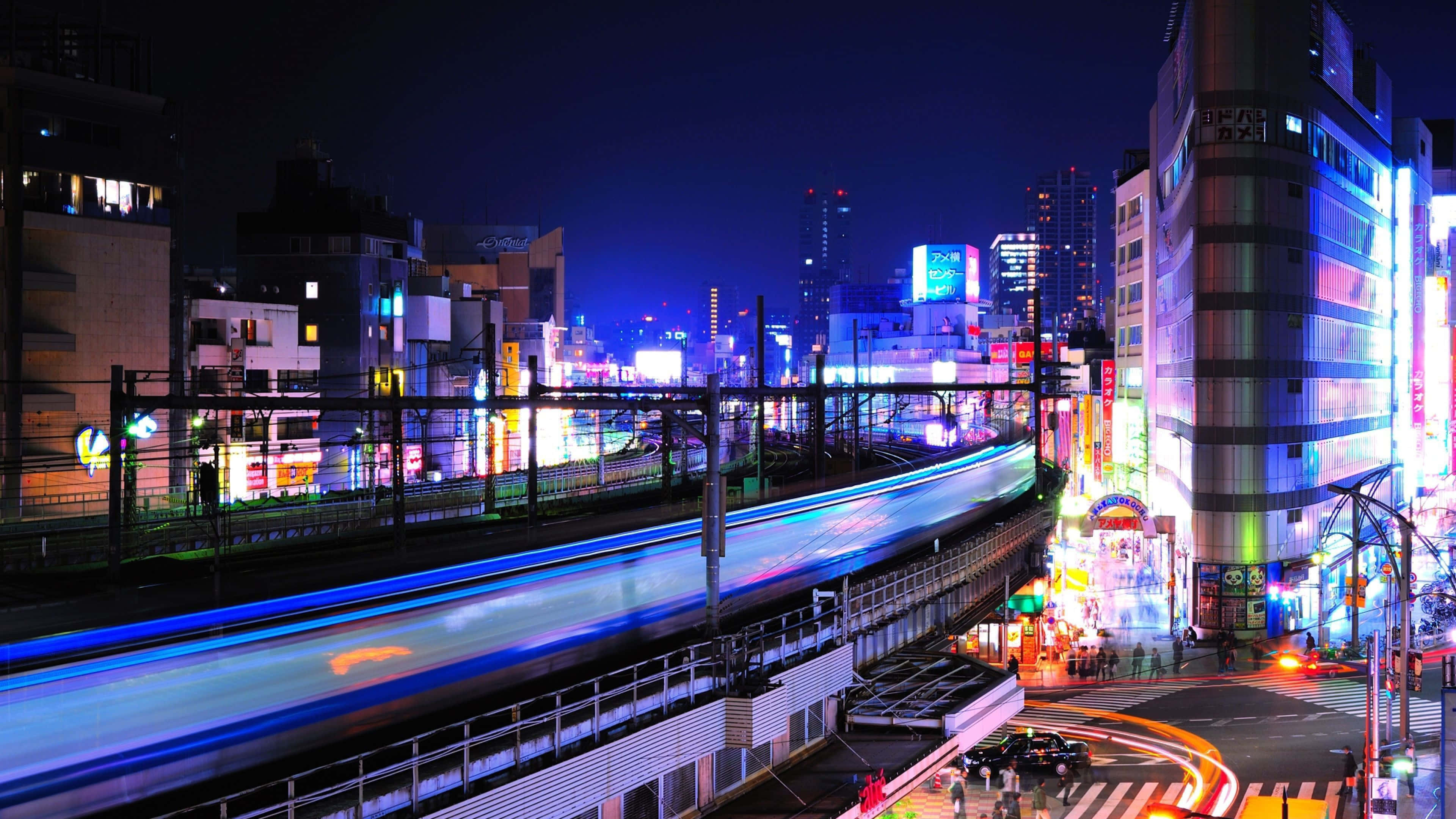 Harajuku 桌布 壁紙 — 充滿活力的夜景城市景觀，閃爍的霓虹燈招牌和移動中的火車及汽車留下的光軌。