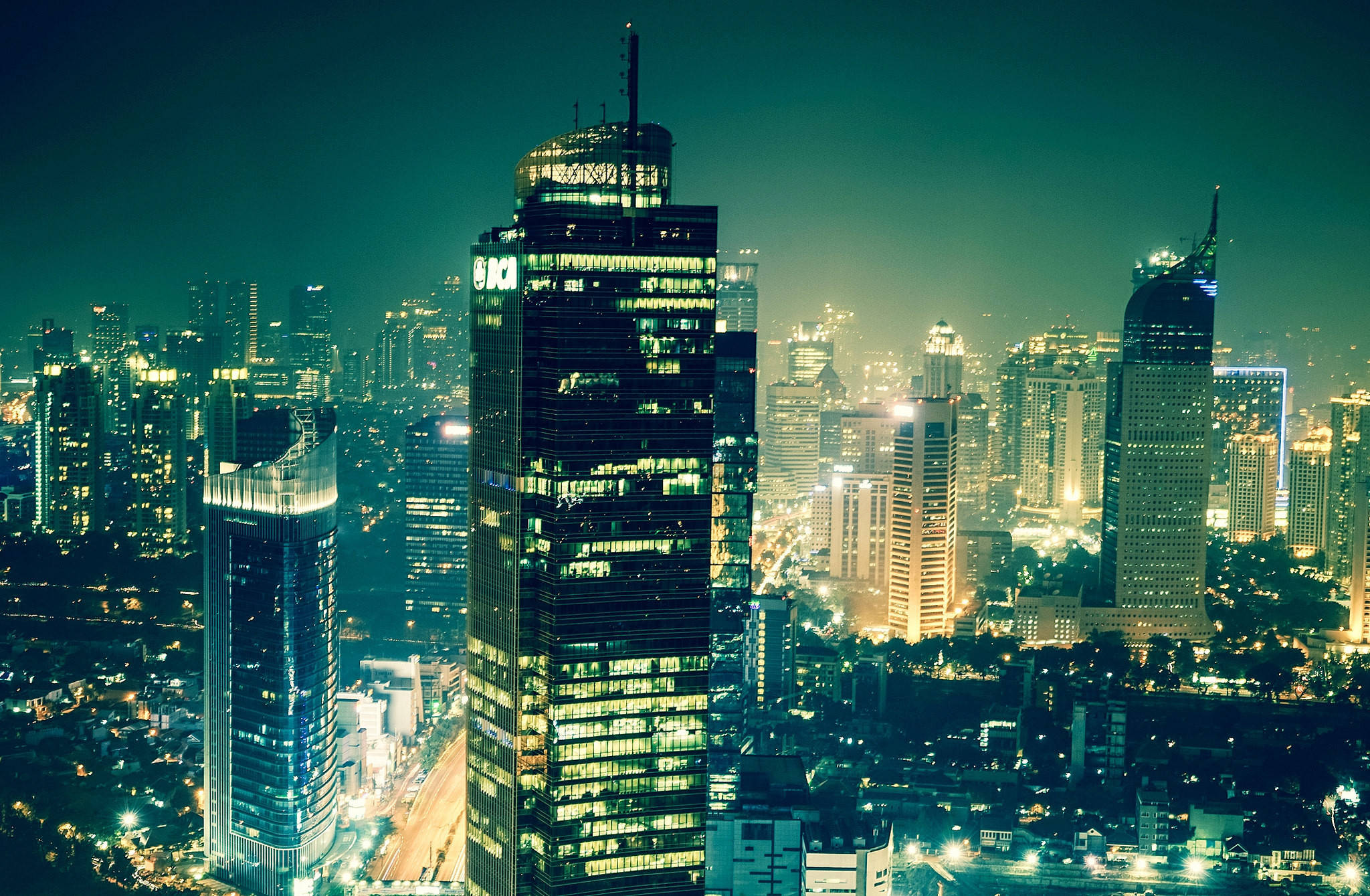 Jakarta 桌布 壁紙 — 充滿活力的夜景城市景觀，高聳的摩天大樓在深藍綠色天空下閃耀。