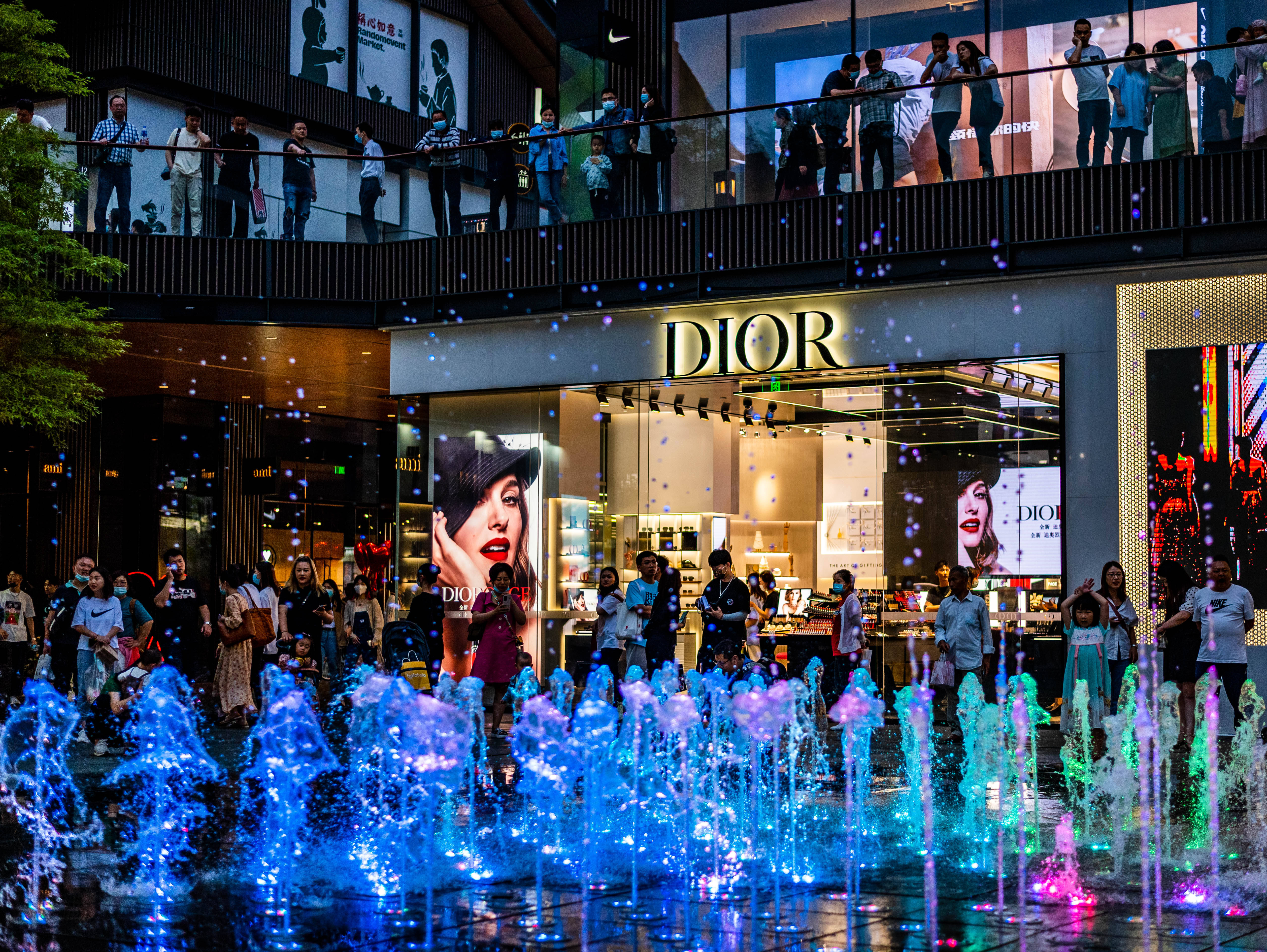 Dior 動態 桌布 壁紙 — 充滿活力的夜間城市景觀，Dior 商店前有發光的藍色和紫色噴泉，周圍是人群和明亮的商店櫥窗。