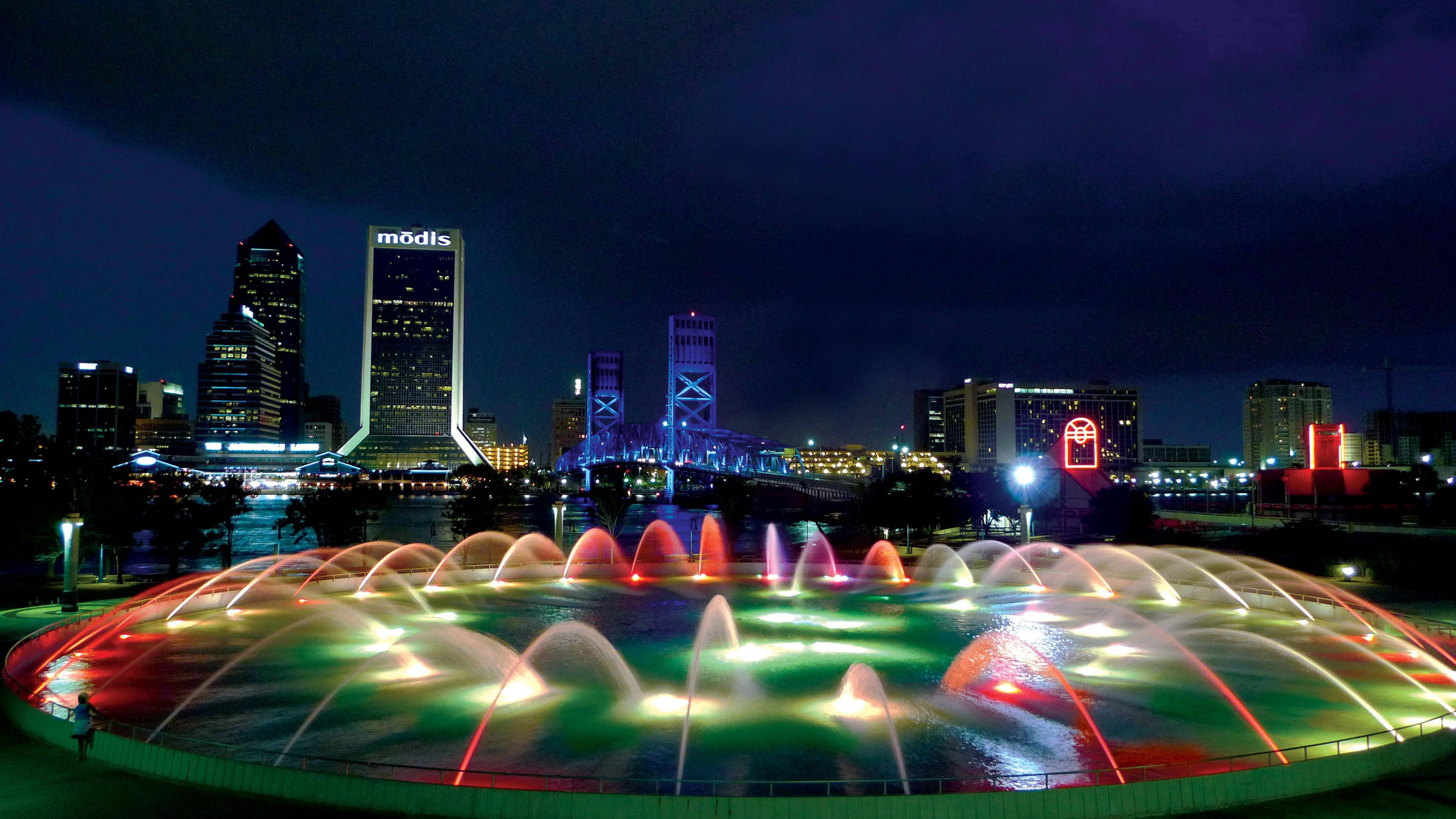 Jacksonville 桌布 壁紙 — 充滿活力的夜間城市景觀，圓形噴泉噴出彩虹色的水柱，在黑暗的天空和發光的摩天大樓襯托下閃耀。