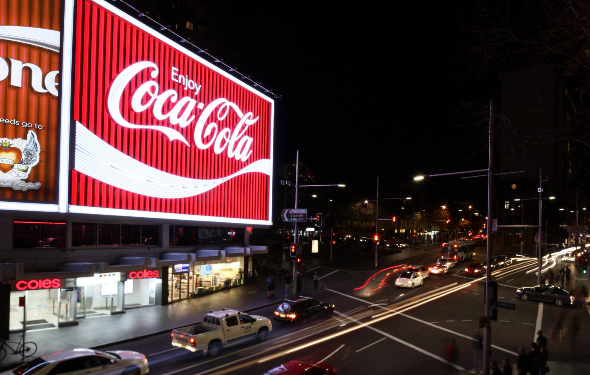 Sydney 桌布 壁紙 — 充滿活力的夜間城市景觀，巨大的發光可口可樂廣告牌照亮了繁忙的街道，伴隨著汽車燈光的流線。