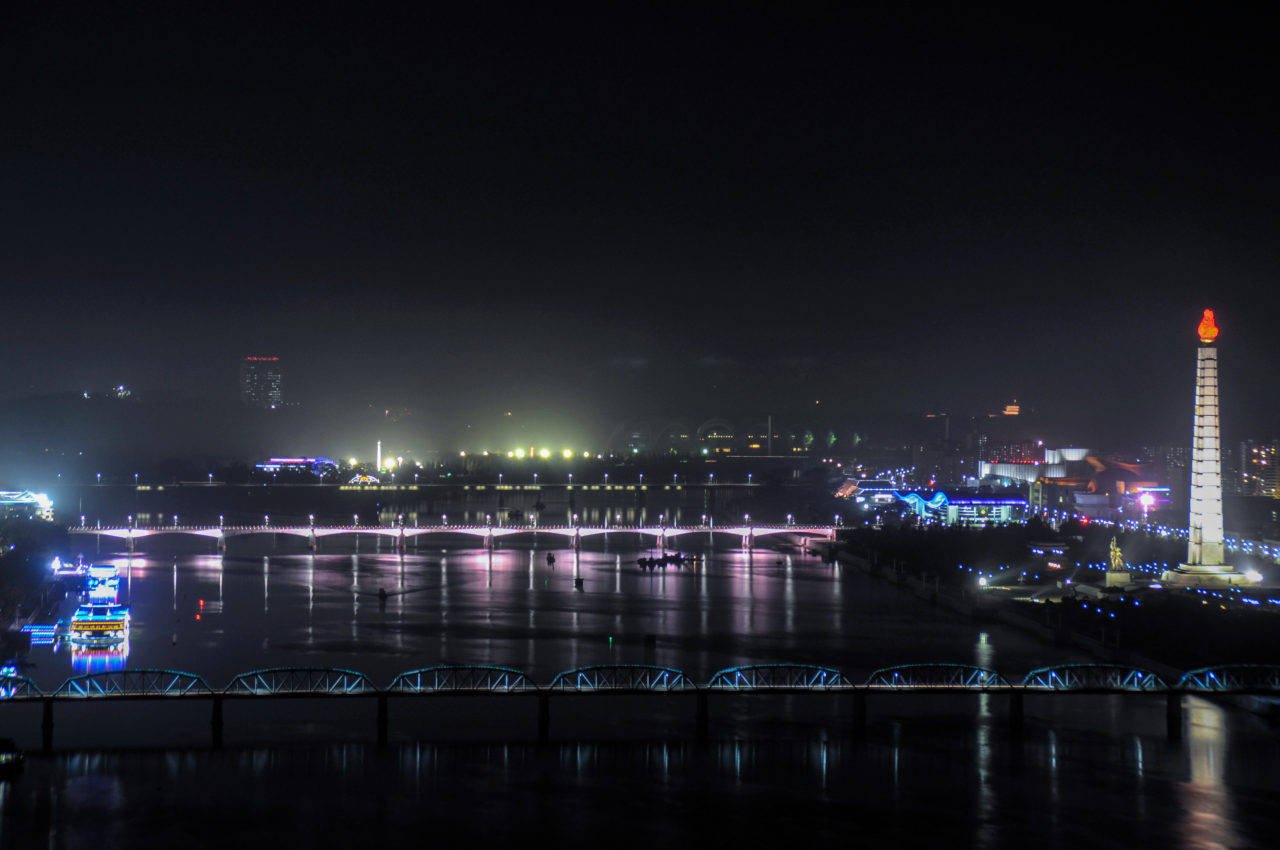 韓國 桌布 壁紙 — 充滿活力的夜間城市景觀，有一座發光的橋樑橫跨水面，以及一個頂端有火焰的高大紀念碑。