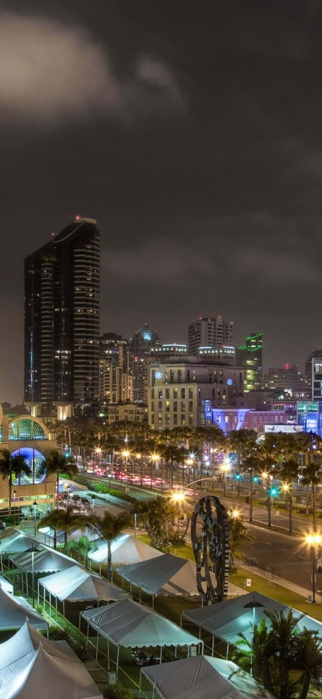 San Diego iPhone 桌布 壁紙 — 充滿活力的夜間城市景觀，有燈火通明的摩天大樓、棕櫚樹和發光的路燈，在陰雲密布的天空下。