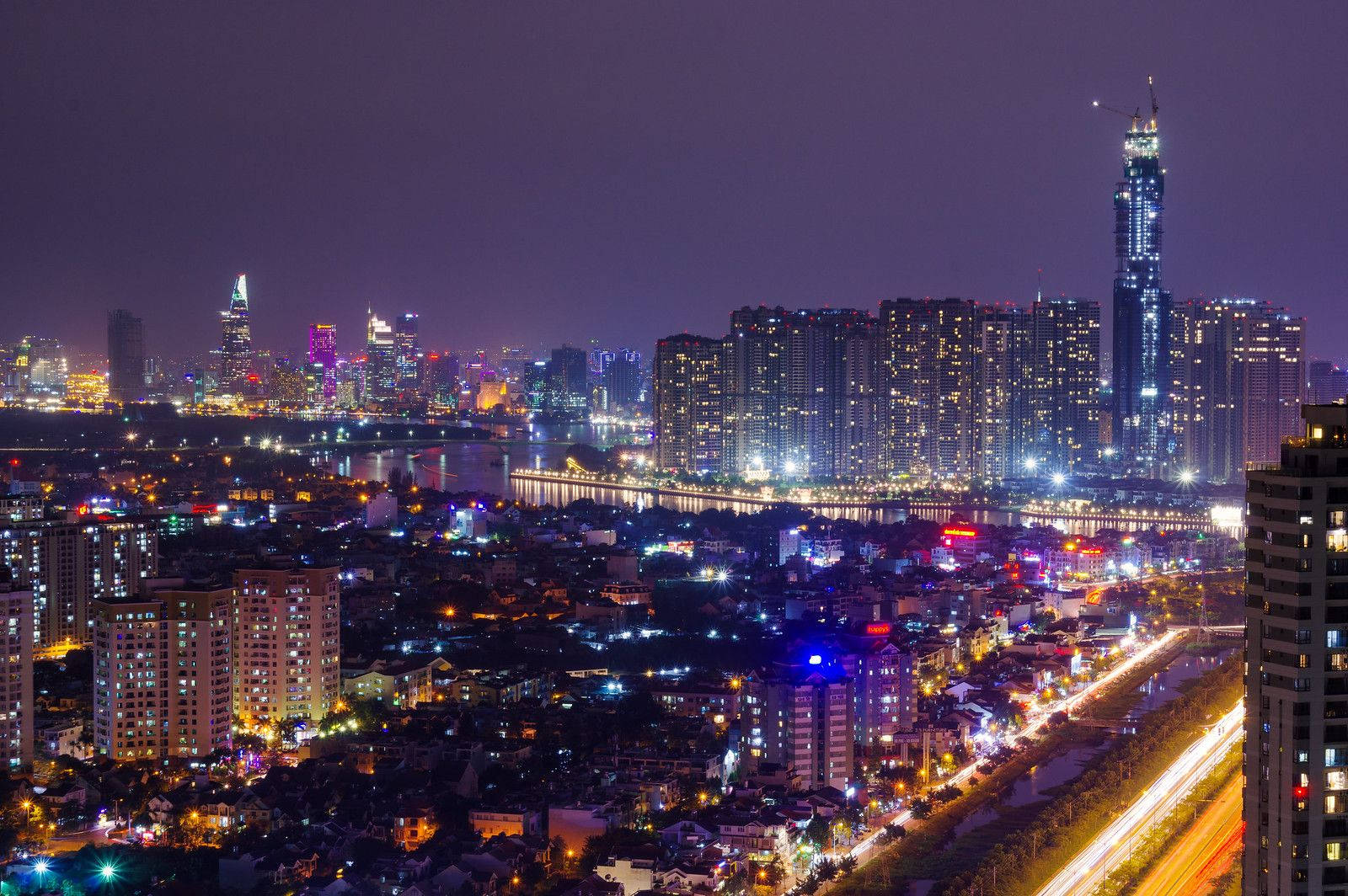 Ho Chi Minh City 桌布 壁紙 — 充滿活力的夜間城市景觀，河邊有發光的摩天大樓和交通燈跡。