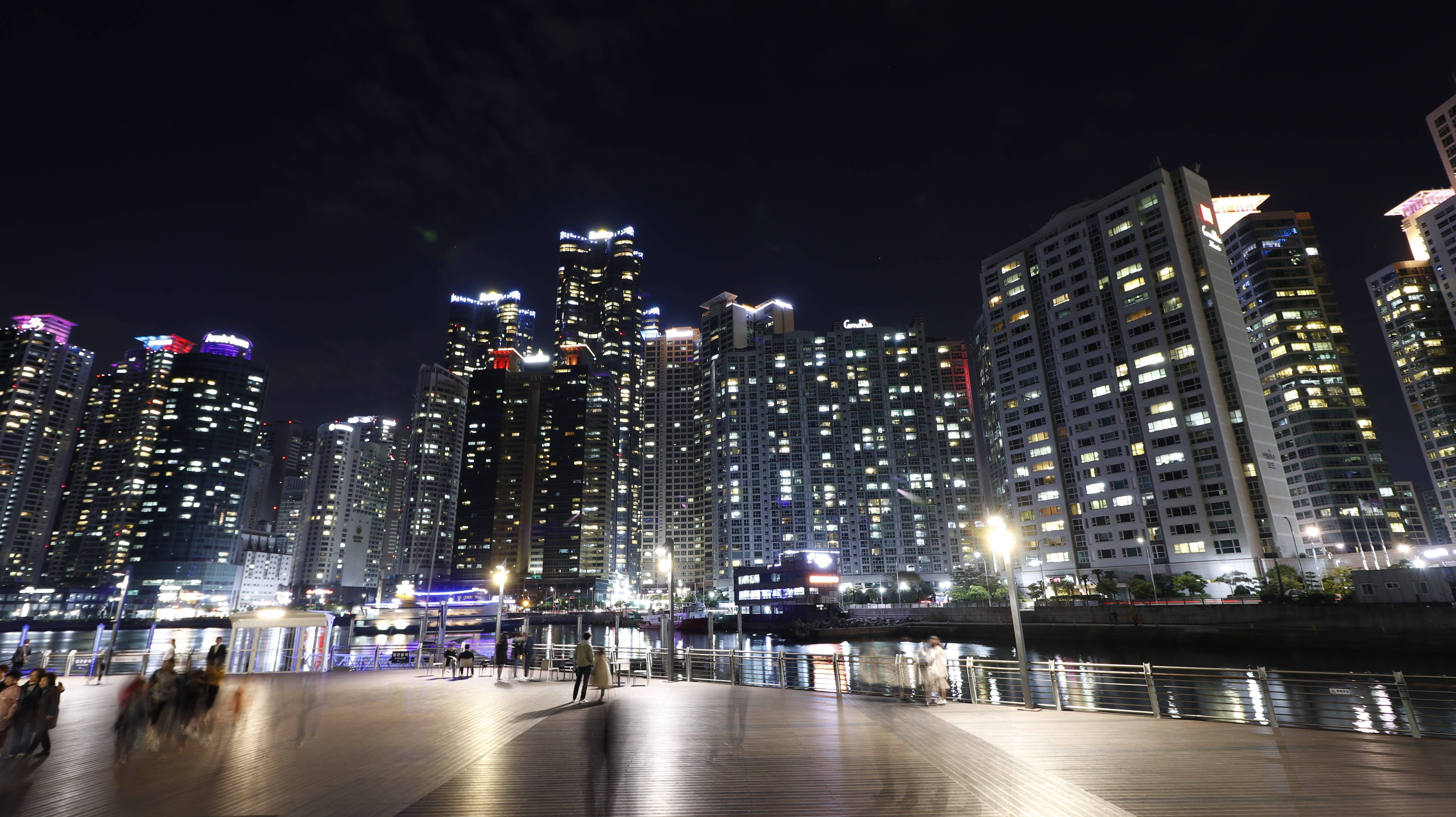 Busan 桌布 壁紙 — 充滿活力的夜間城市景觀，燈火通明的摩天大樓倒映在水面上，木質長廊上模糊的行人。