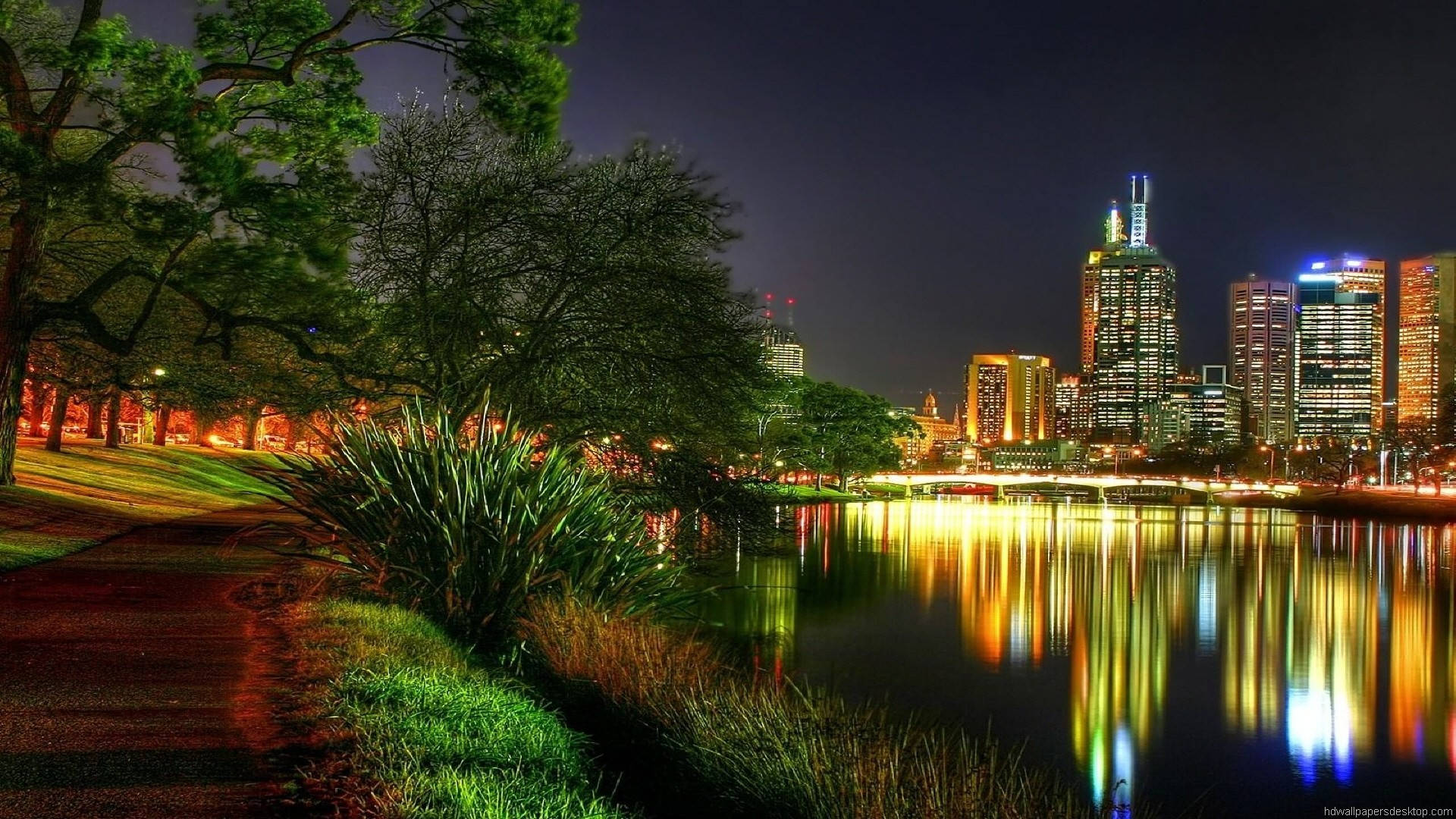 1920x1080 高畫質自然 桌布 壁紙 — 充滿活力的夜間城市景觀，發光的摩天大樓倒映在平靜的湖面上，周圍環繞著茂密的綠樹和蜿蜒的小徑。