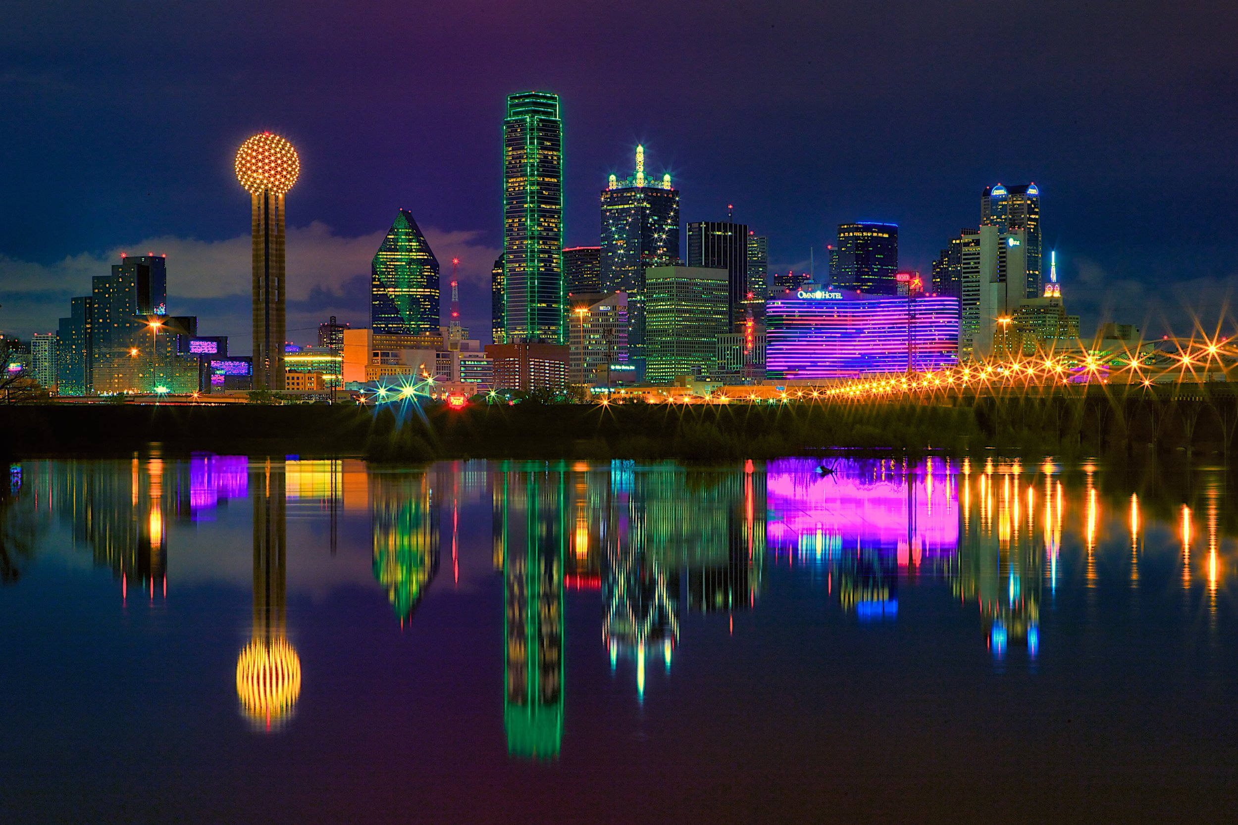 德州 桌布 壁紙 — 充滿活力的夜間城市景觀，色彩繽紛的摩天大樓和發光的球體塔倒映在平靜的水面上。