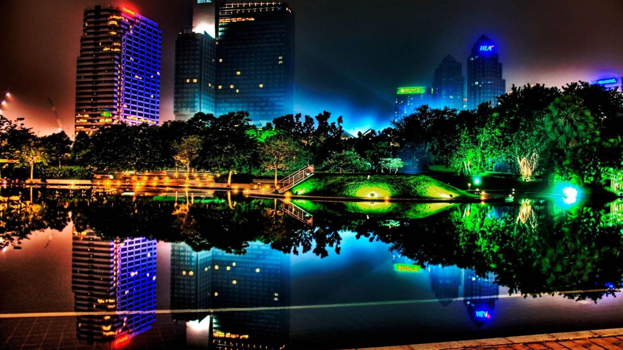 霓虹城市動態圖 桌布 壁紙 — 充滿活力的夜間城市景觀，霓虹燈照亮的摩天大樓倒映在平靜的池塘中，周圍環繞著發光的綠色樹木。