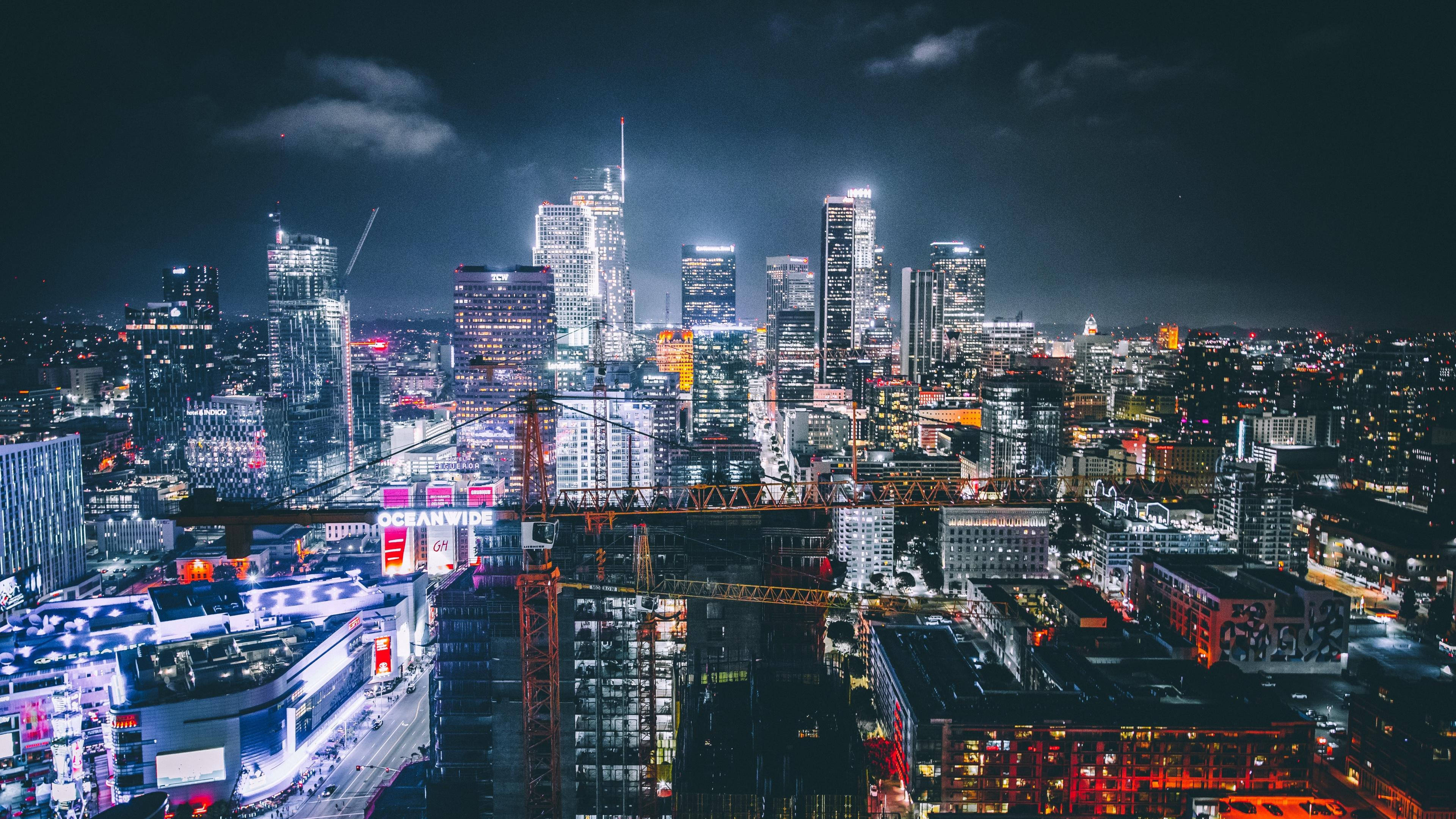 洛杉磯 4K 桌布 壁紙 — 充滿活力的夜間城市景觀，高聳的摩天大樓在黑暗的天空下閃耀，一座橋樑橫跨城市區域，建築起重機在燈光中呈現剪影。