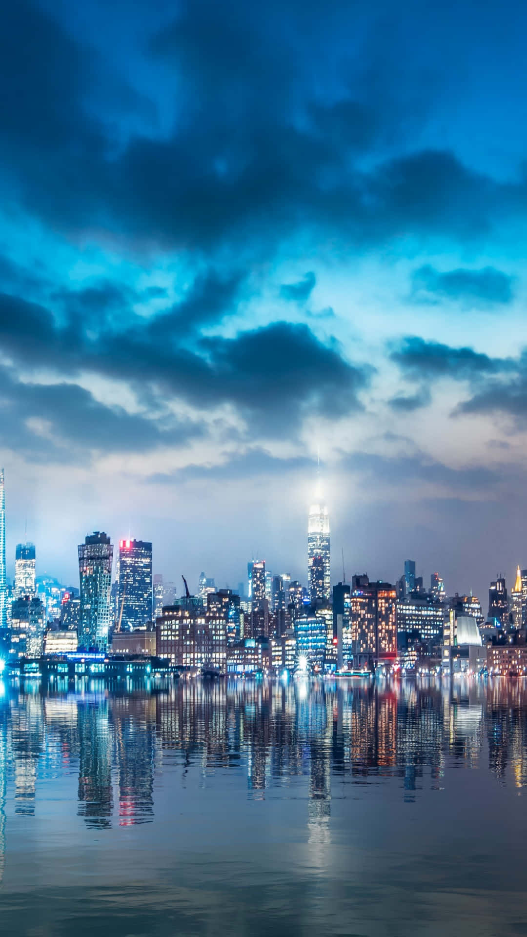 城市風景 桌布 壁紙 — 充滿活力的夜間城市景觀，高聳的摩天大樓在平靜的水面上倒映，天空憂鬱藍色。