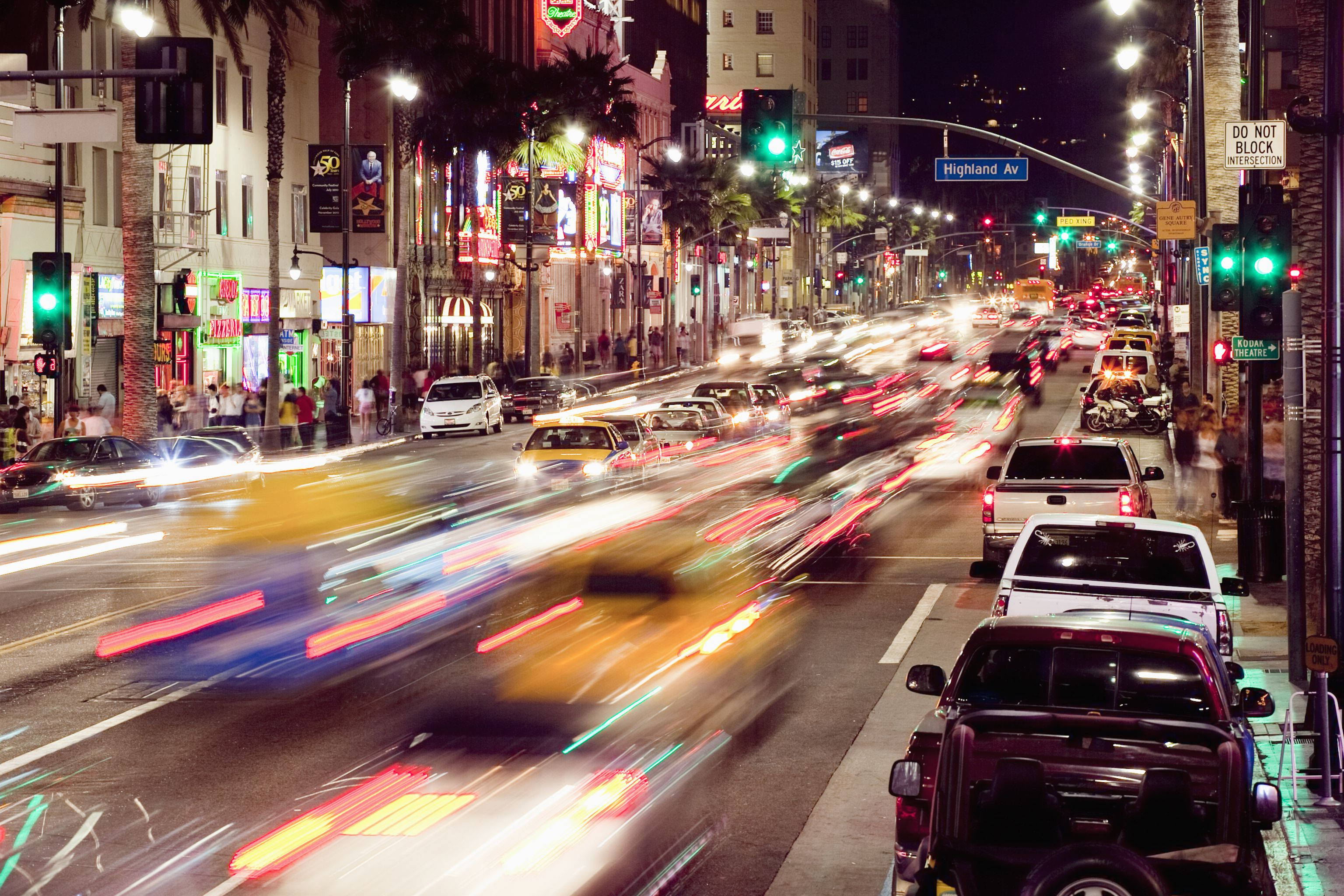 hollywood street 桌布 壁紙 — 充滿活力的夜間城市街道，行駛中的汽車留下光跡，閃爍的霓虹燈照亮棕櫚樹和行人。