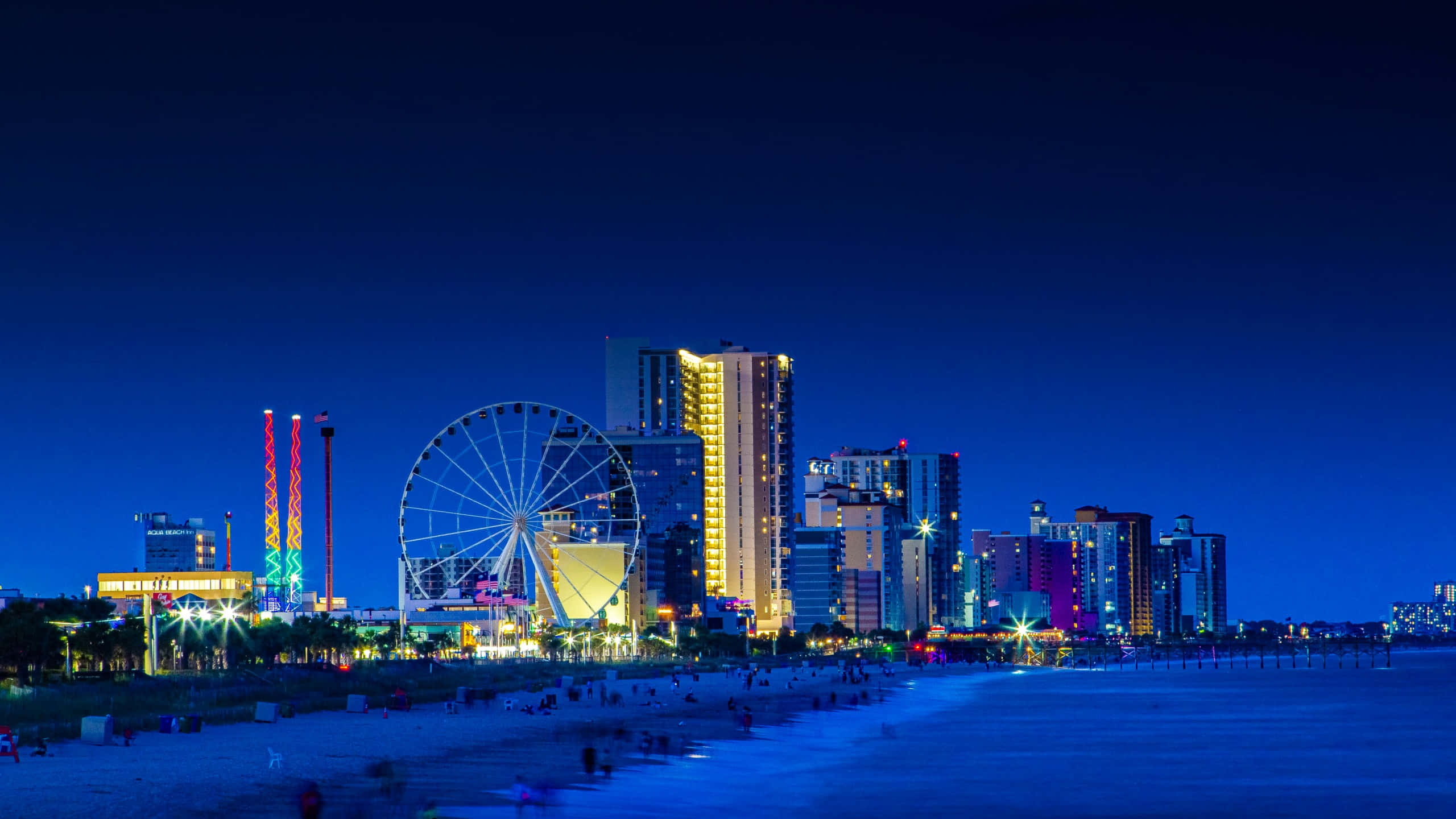 Myrtle Beach 桌布 壁紙 — 充滿活力的夜間海岸城市景觀，閃爍的摩天輪和色彩繽紛的建築物照亮了沙灘上方的深藍色天空。