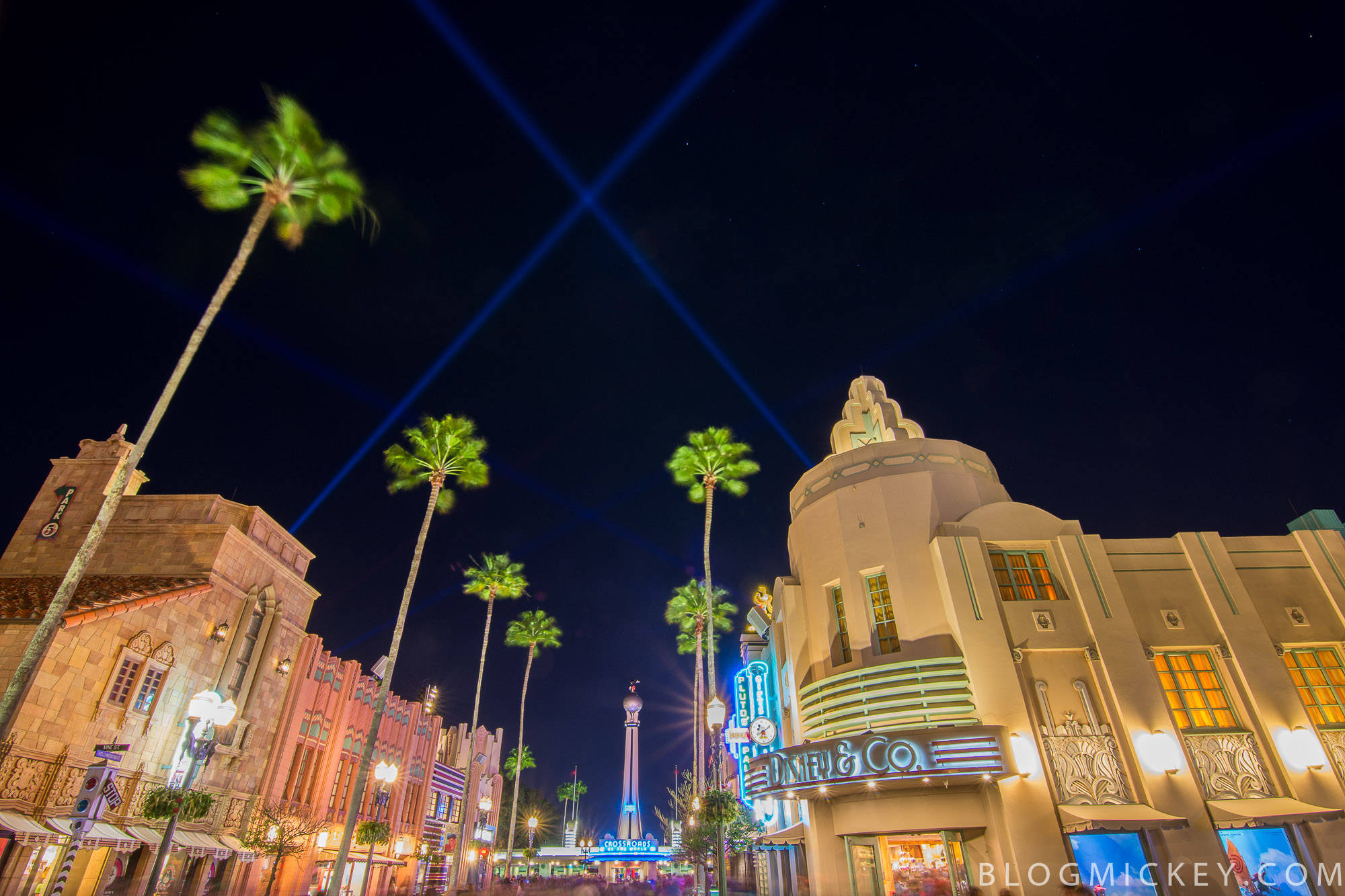 hollywood street 桌布 壁紙 — 充滿活力的夜間街景，棕櫚樹和裝飾藝術風格的建築被五彩繽紛的燈光和劃過天空的藍色雷射光束照亮。