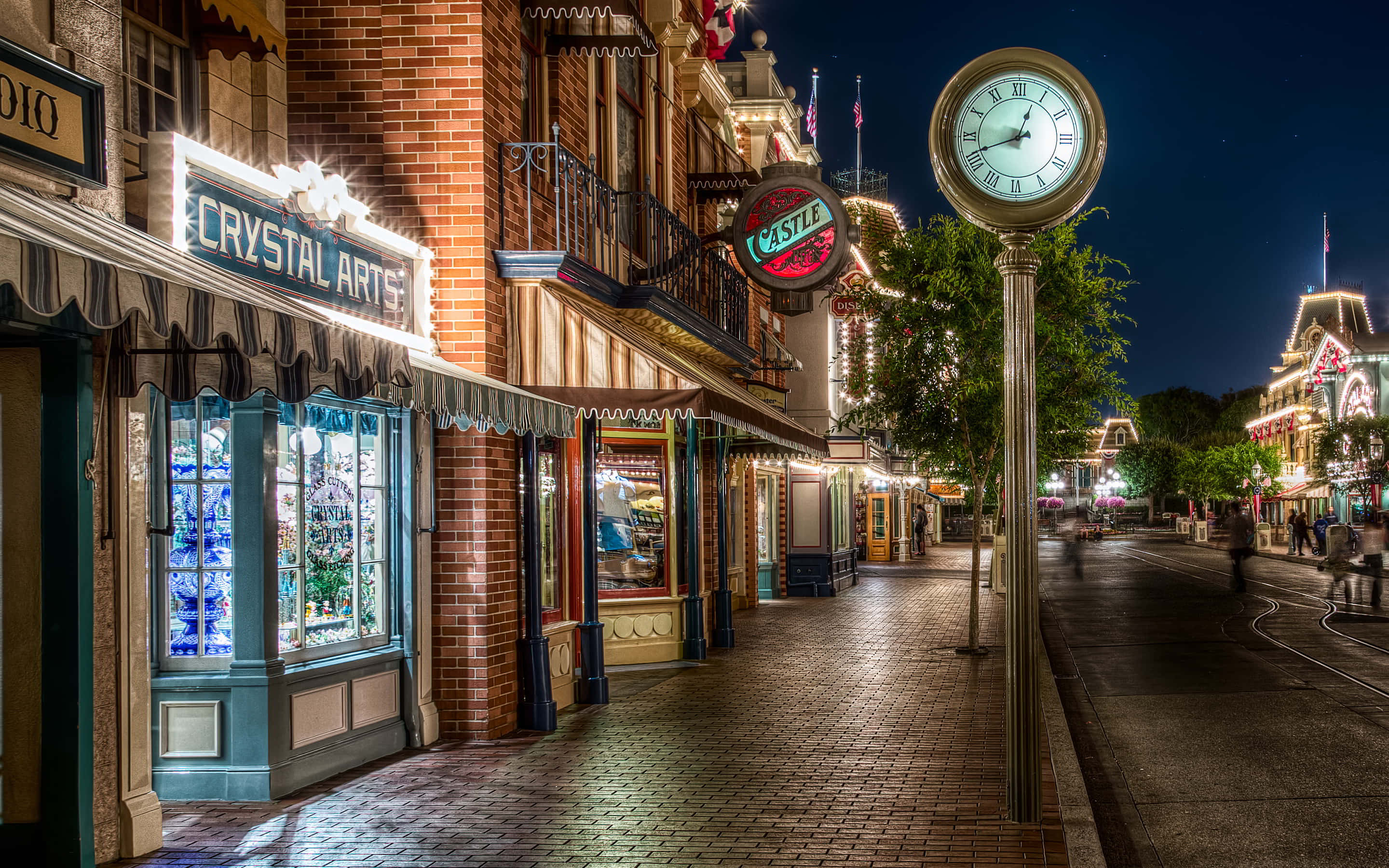 Disneyland 桌布 壁紙 — 充滿活力的夜間街景，閃爍的霓虹燈招牌和一個大時鐘照亮了磚砌的人行道。