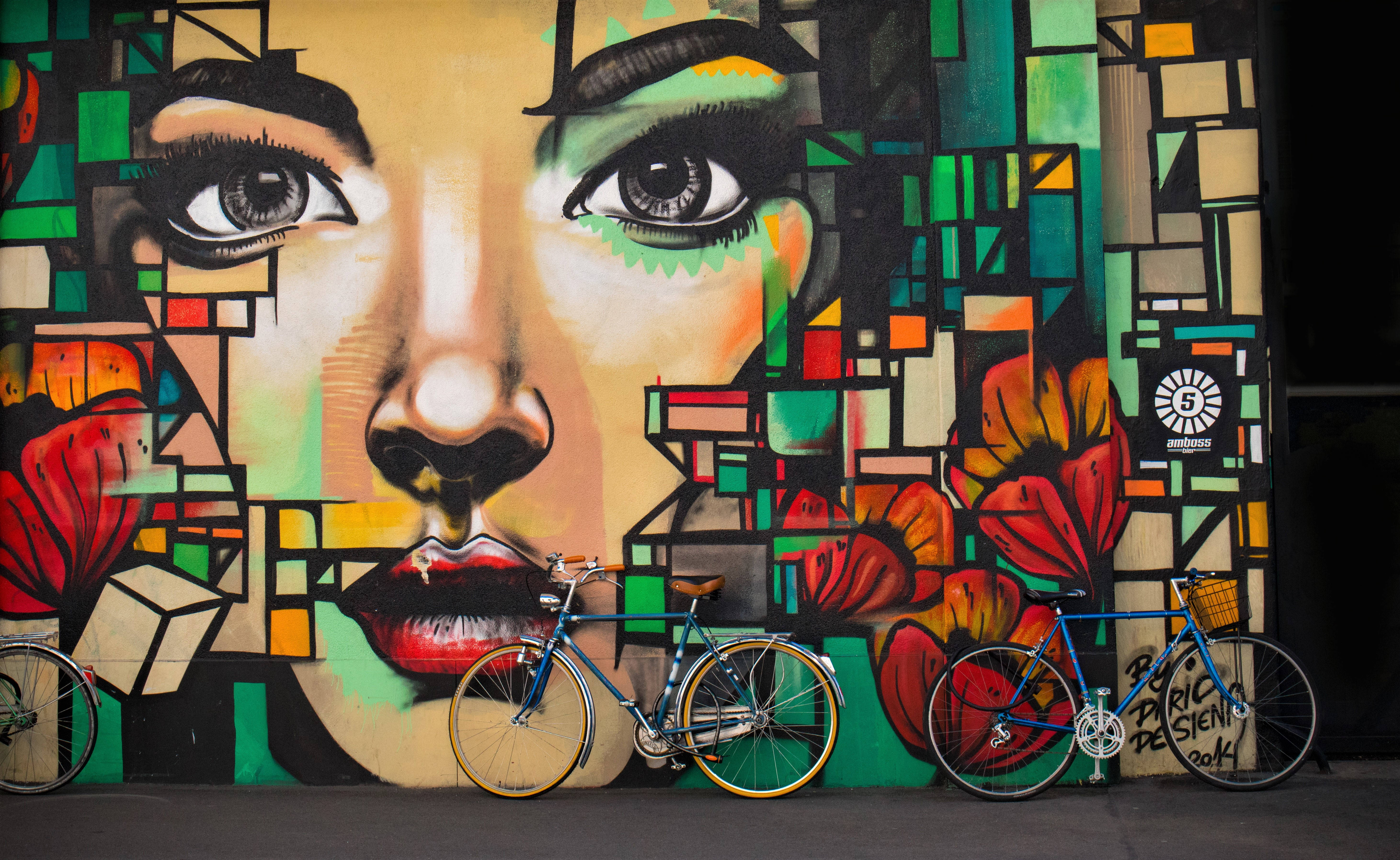 Bicycle 桌布 壁紙 — 充滿活力的幾何塗鴉壁畫，描繪一位女性臉龐，帶有鮮豔的紅唇和花朵，前方停著兩輛藍色腳踏車作為框架。