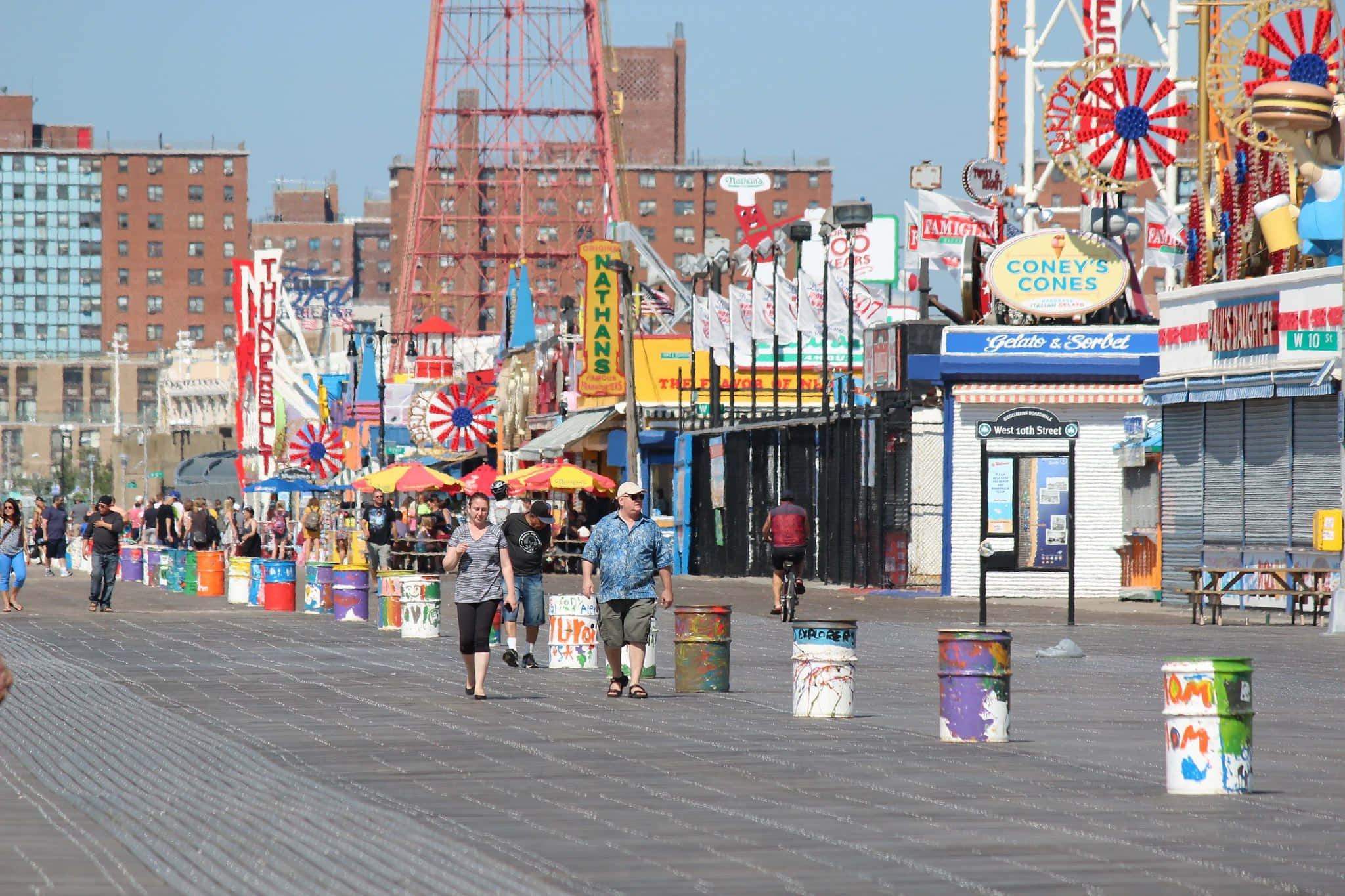 Boardwalk 桌布 壁紙 — 充滿活力的康尼島木板路，在明媚陽光下擠滿人群，色彩繽紛的嘉年華遊樂設施和彩繪桶排列在路邊。