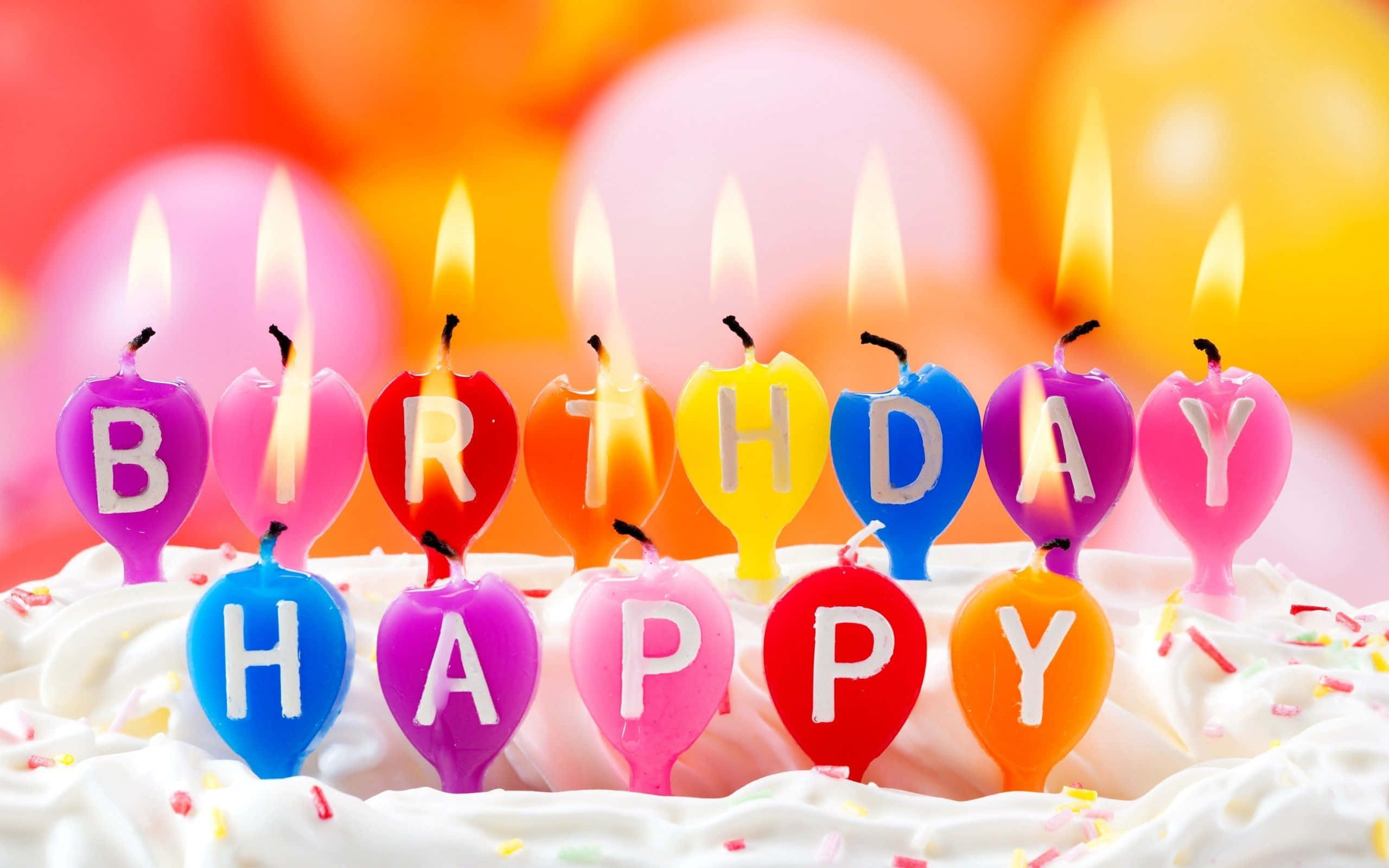 Birthday Wishes 桌布 壁紙 — 充滿活力的心形生日蠟燭拼出「HAPPY BIRTHDAY」，在鋪滿彩色糖霜的蛋糕上閃耀，背景是模糊的氣球。