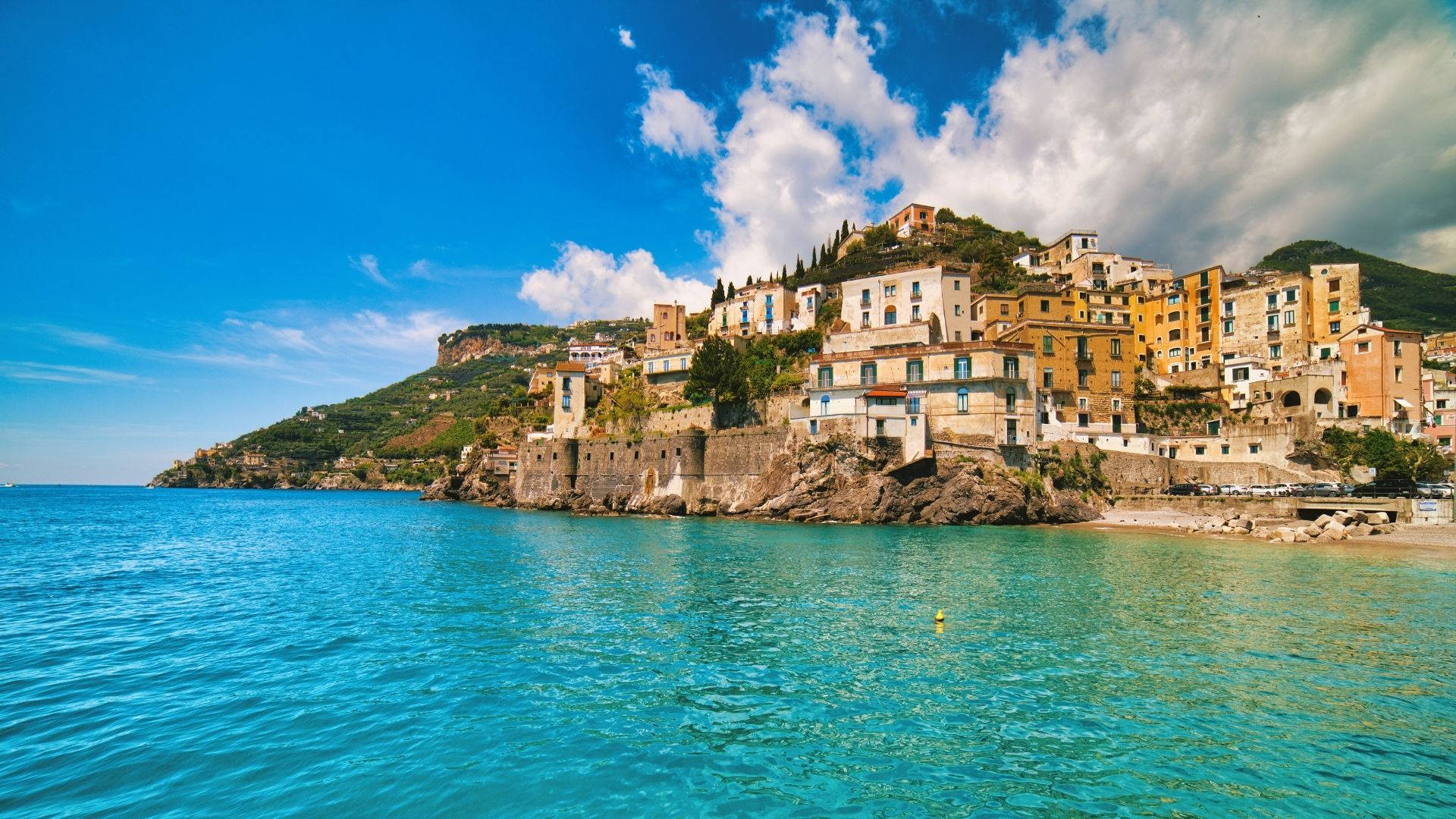 Amalfi Coast 桌布 壁紙 — 充滿活力的懸崖村莊，赤陶屋頂建築矗立在碧綠海水之上，藍天白雲下。