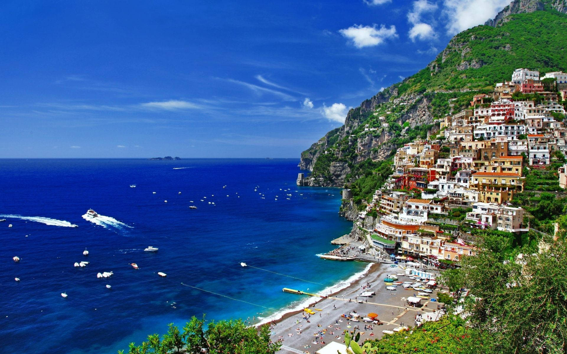 Capri 桌布 壁紙 — 充滿活力的懸崖村莊波西塔諾依附在陡峭的綠色山丘上，俯瞰著點綴著船隻的碧綠大海，上方是明亮的藍天。