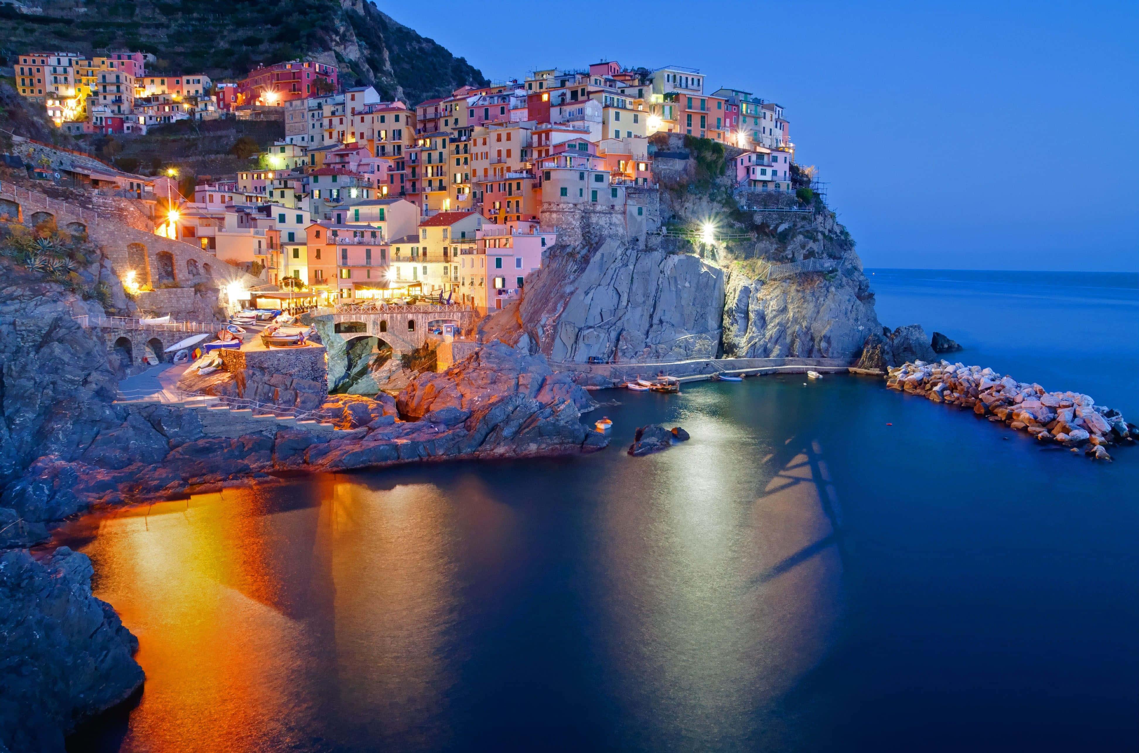 Italy Aesthetic 桌布 壁紙 — 充滿活力的懸崖村莊燈光在暮色中溫暖地倒映在平靜的藍色水面上。