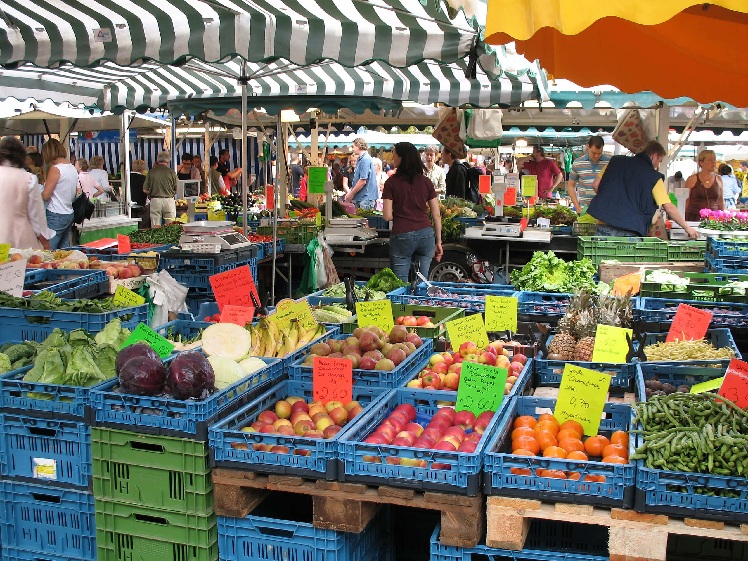 Farmers Market 桌布 壁紙 — 充滿活力的戶外市場攤位在條紋遮陽篷下堆滿了色彩繽紛的新鮮農產品，購物者正在瀏覽。