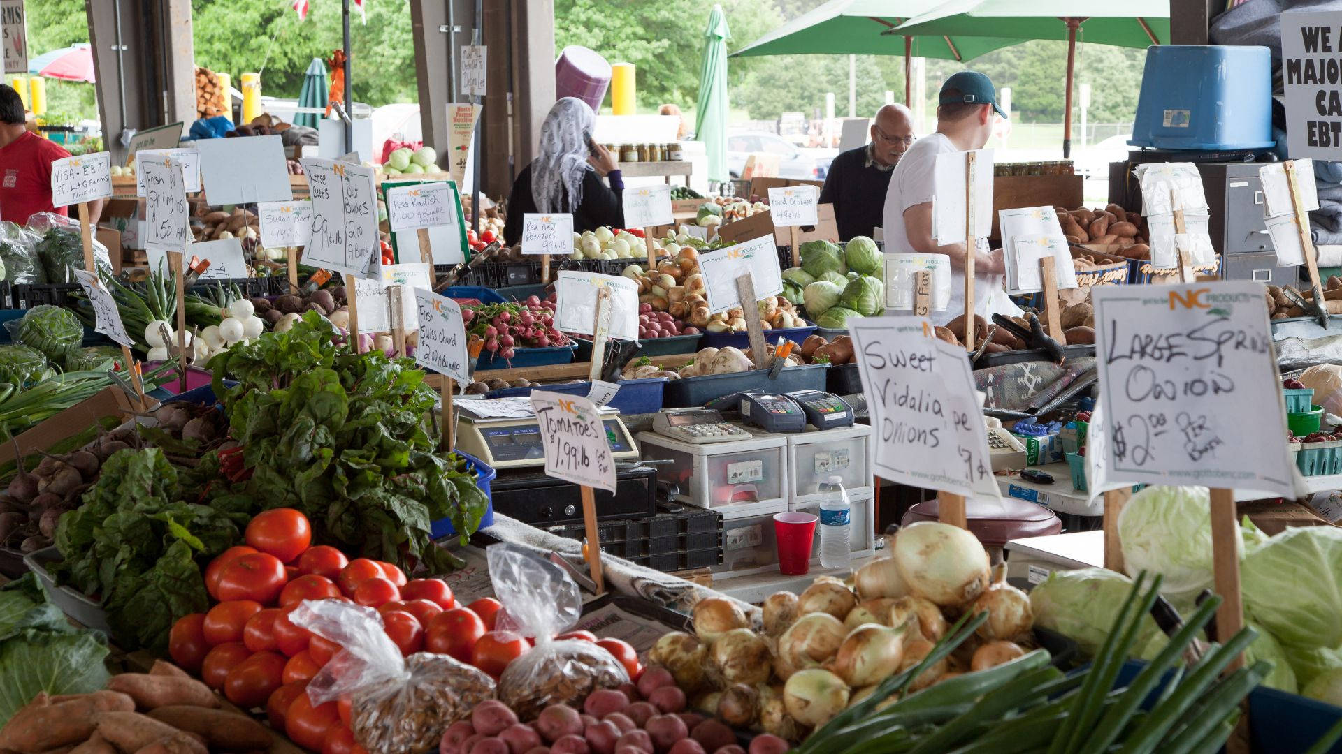 Vegetables 桌布 壁紙 — 充滿活力的戶外農貿市場攤位，在綠色遮陽傘下堆滿了色彩繽紛的新鮮農產品，如番茄、洋蔥和高麗菜。