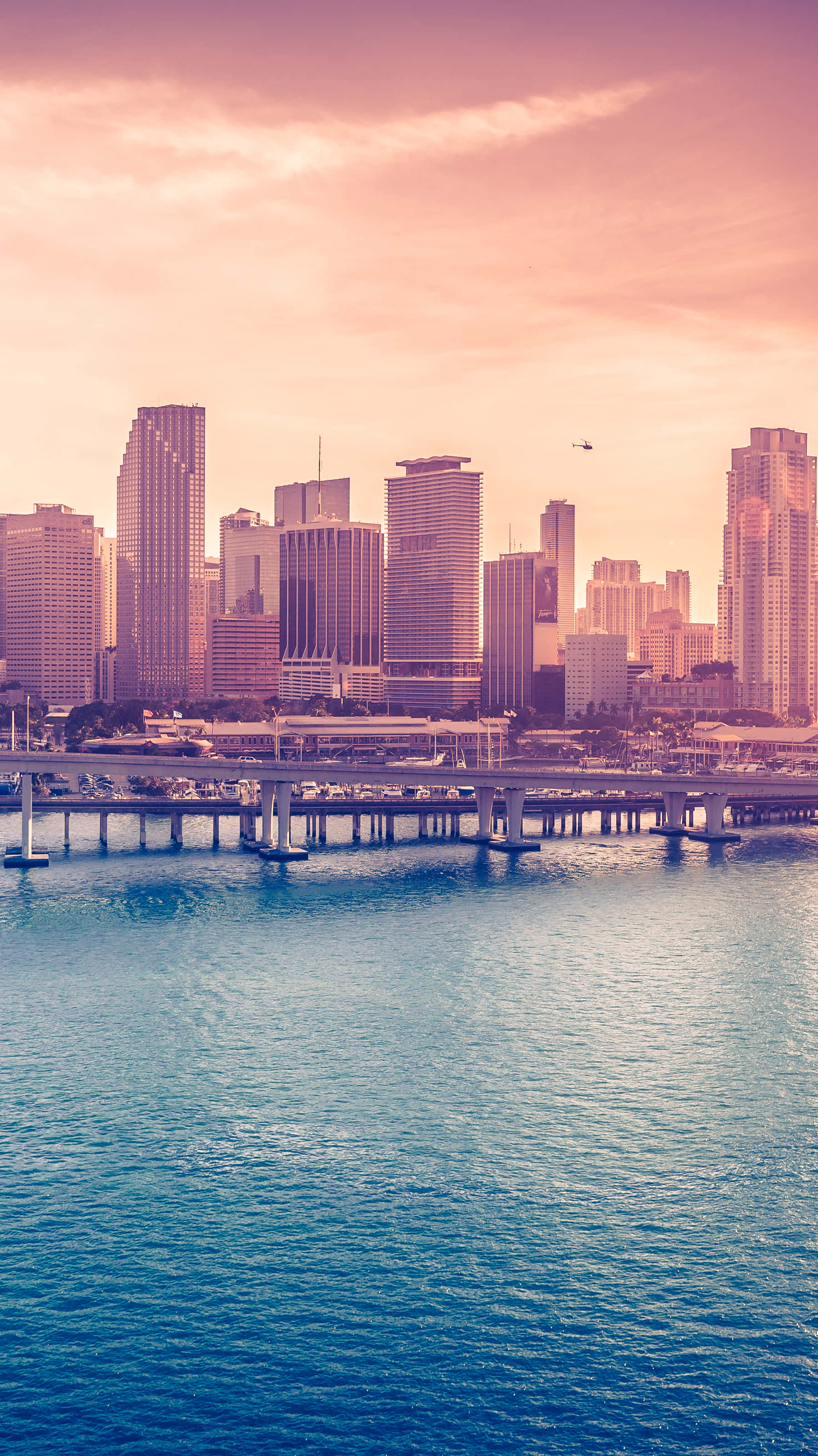 Miami 桌布 壁紙 — 充滿活力的日落以溫暖的粉紅色和橙色色調沐浴著現代城市的天際線，並倒映在下方平靜的藍色水面上。