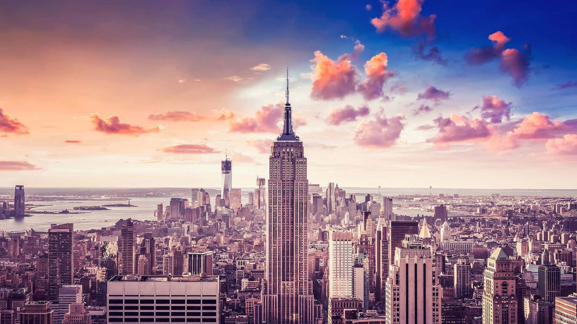 New York 桌布 壁紙 — 充滿活力的日落以溫暖的粉紅色和橙色色調沐浴著標誌性的紐約市天際線，帝國大廈聳立在密集的城市景觀之上。