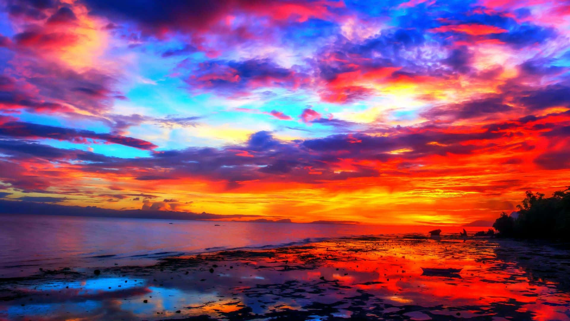 Colorful Sky 桌布 壁紙 — 充滿活力的日落將天空和海水染成火紅、橙色和電光藍，映照在寧靜的海灘上。