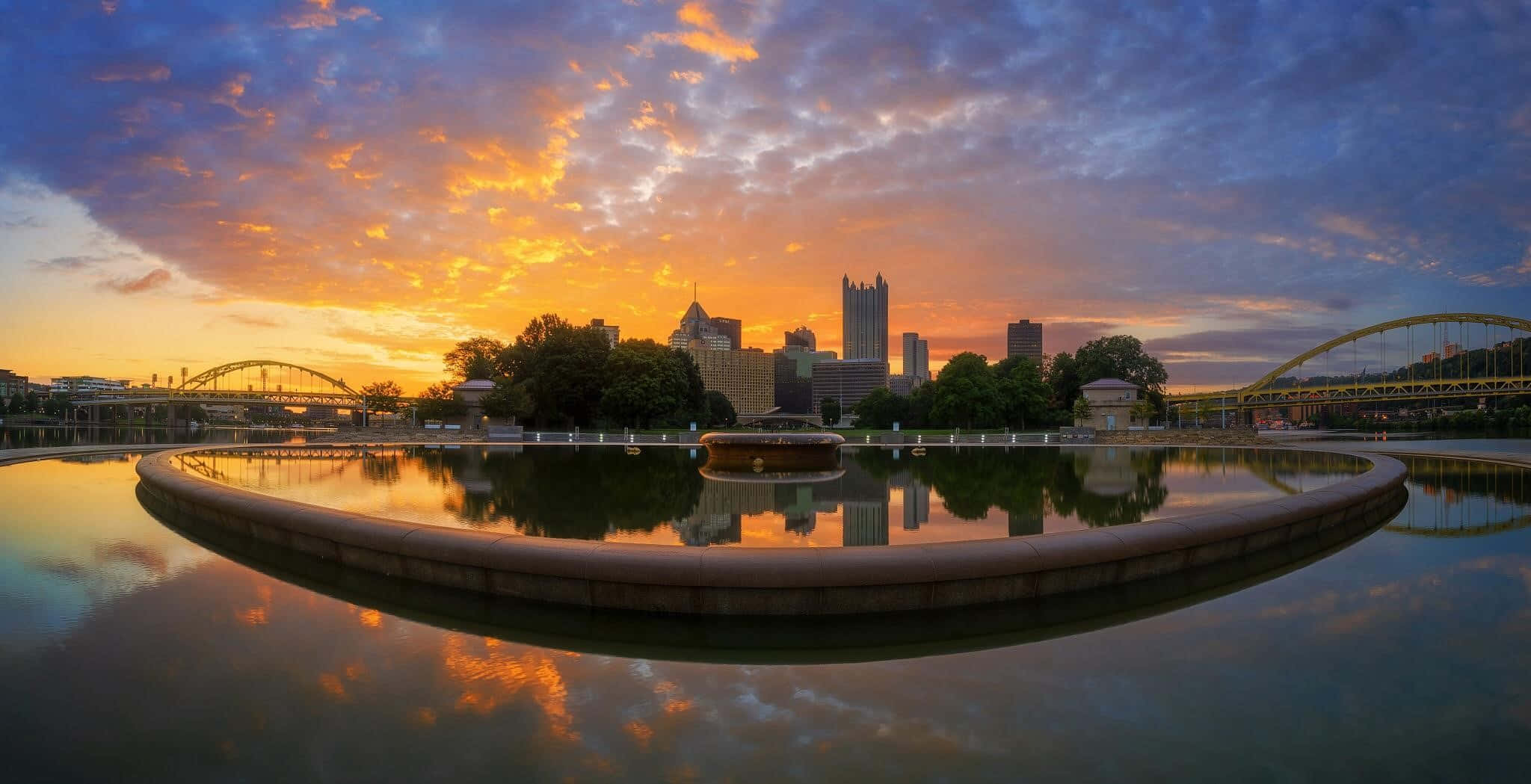 Pittsburgh Skyline 桌布 壁紙 — 充滿活力的日落將天空染成橙色和紫色，完美地倒映在平靜的圓形噴泉中，背景是城市天際線和橋樑。