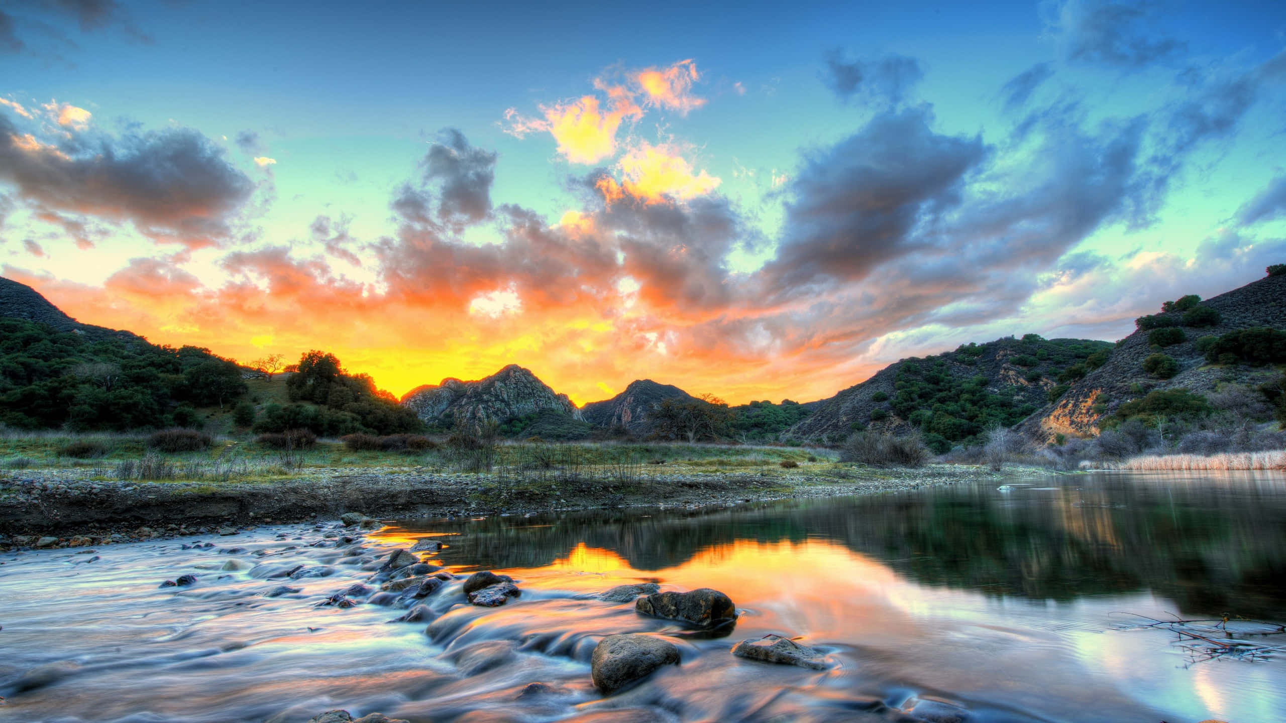 Nature Landscape 桌布 壁紙 — 充滿活力的日落將天空染成火橙色和青綠色的雲彩，倒映在流過岩石的平靜河流中。