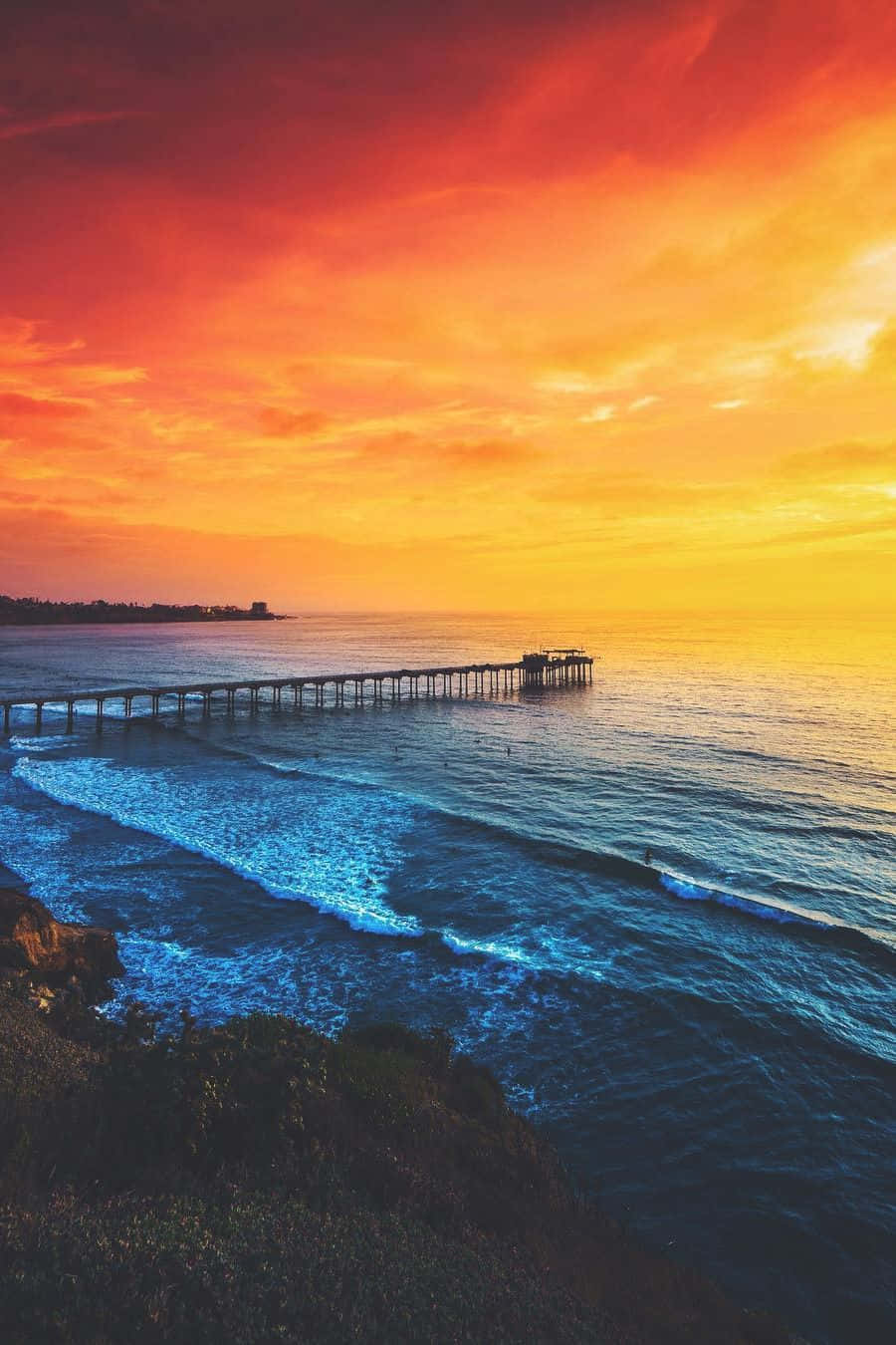 San Diego iPhone 桌布 壁紙 — 充滿活力的日落將天空染成火熱的橙色和紅色，海浪在長碼頭下拍打岩石海岸。